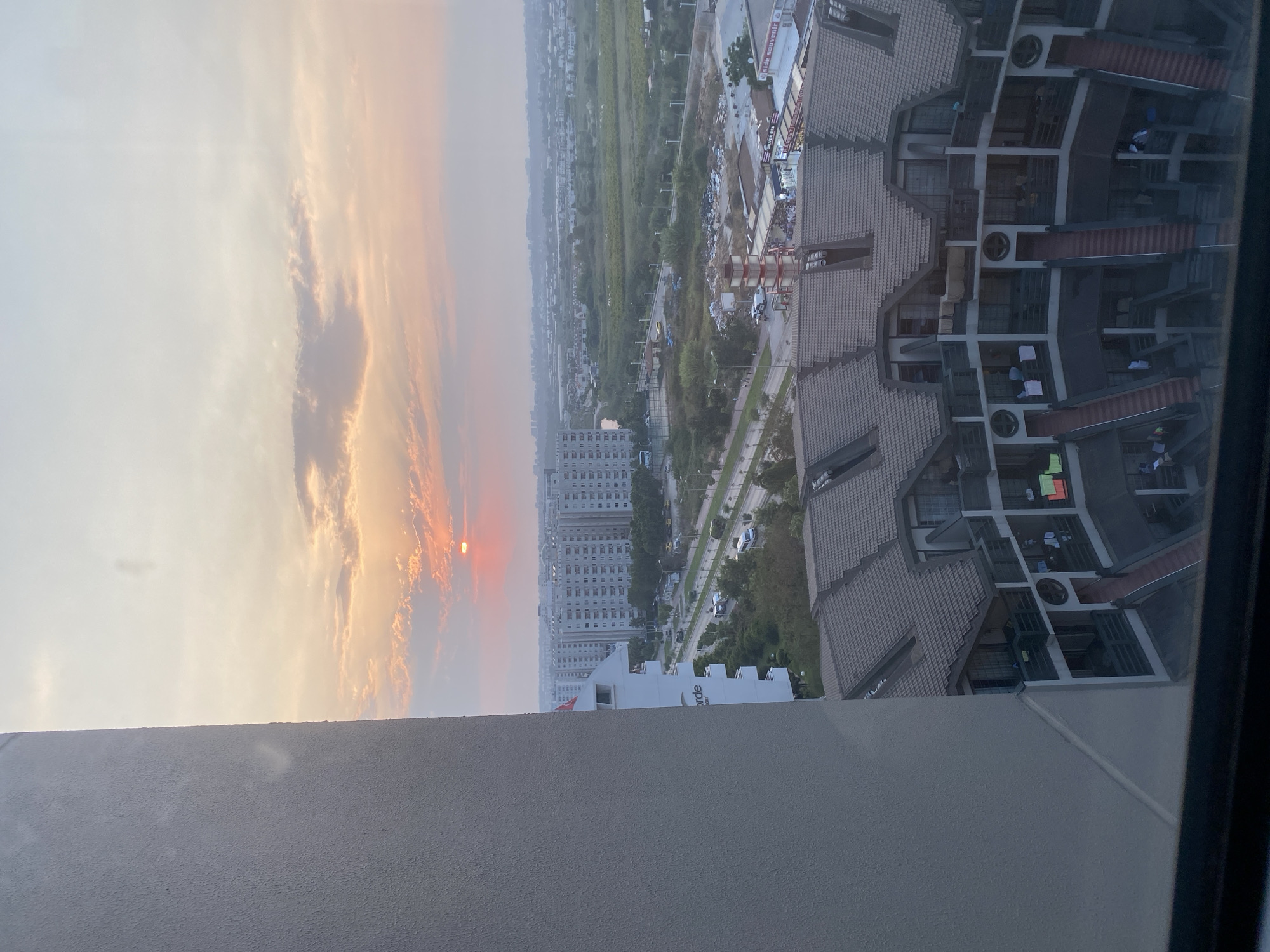 The image captures a sunset over the city of Aksu, Turkey. The foreground is dominated by the side of a large, light-colored building. In the middle ground, a curved building with a tiled roof and multiple balconies is visible, with outdoor stairs leading to different levels. Beyond this, a street runs parallel to a row of tall apartment buildings. In the distance, there are more buildings and agricultural fields, with the hazy horizon indicating the edge of the city. The sun is a small, bright orange disc low in the sky, casting a warm glow through the clouds. There are no discernible people or specific actions taking place. The environment appears to be a suburban or semi-urban area.