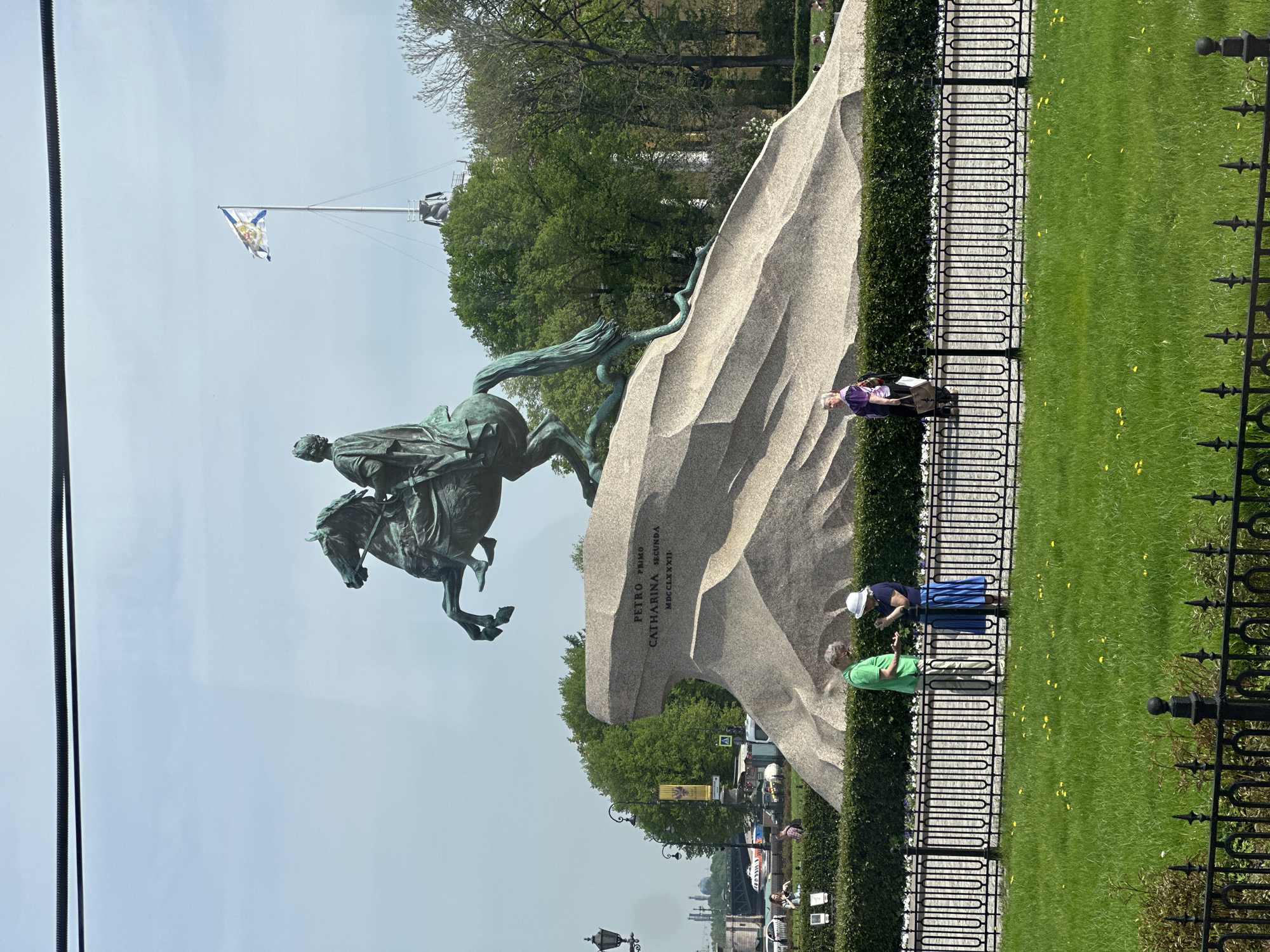 The image shows the Bronze Horseman statue in Saint Petersburg, Russia. The statue depicts Peter the Great on horseback. It is situated on a large granite pedestal. Three people are standing nearby, looking at the statue. A fence runs along the edge of a grassy area. A flag is visible in the background.