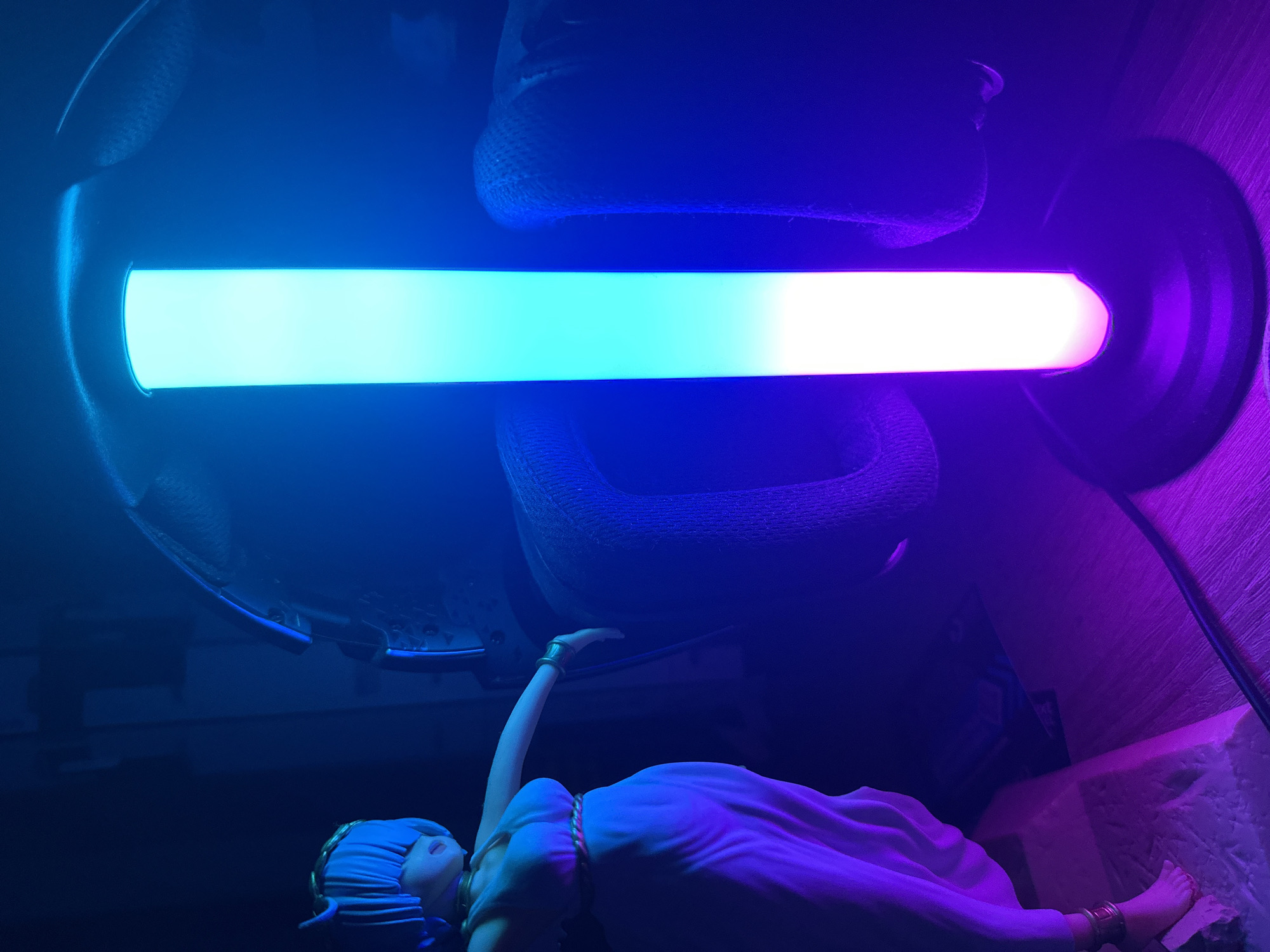 In a dimly lit interior, possibly an apartment or studio, an anime figurine with blue hair and a flowing white dress stands to the left, adorned with gold jewelry. To its right, a cylindrical LED lamp emits a gradient of light, transitioning from white at the bottom to cyan and then a deep blue at the top. The lamp is positioned behind a pair of large, dark-colored headphones. The floor appears to be wooden and is illuminated by the ambient purple and blue light from the lamp. The overall atmosphere is dark and moody, with the vibrant colors of the lamp creating a striking contrast.