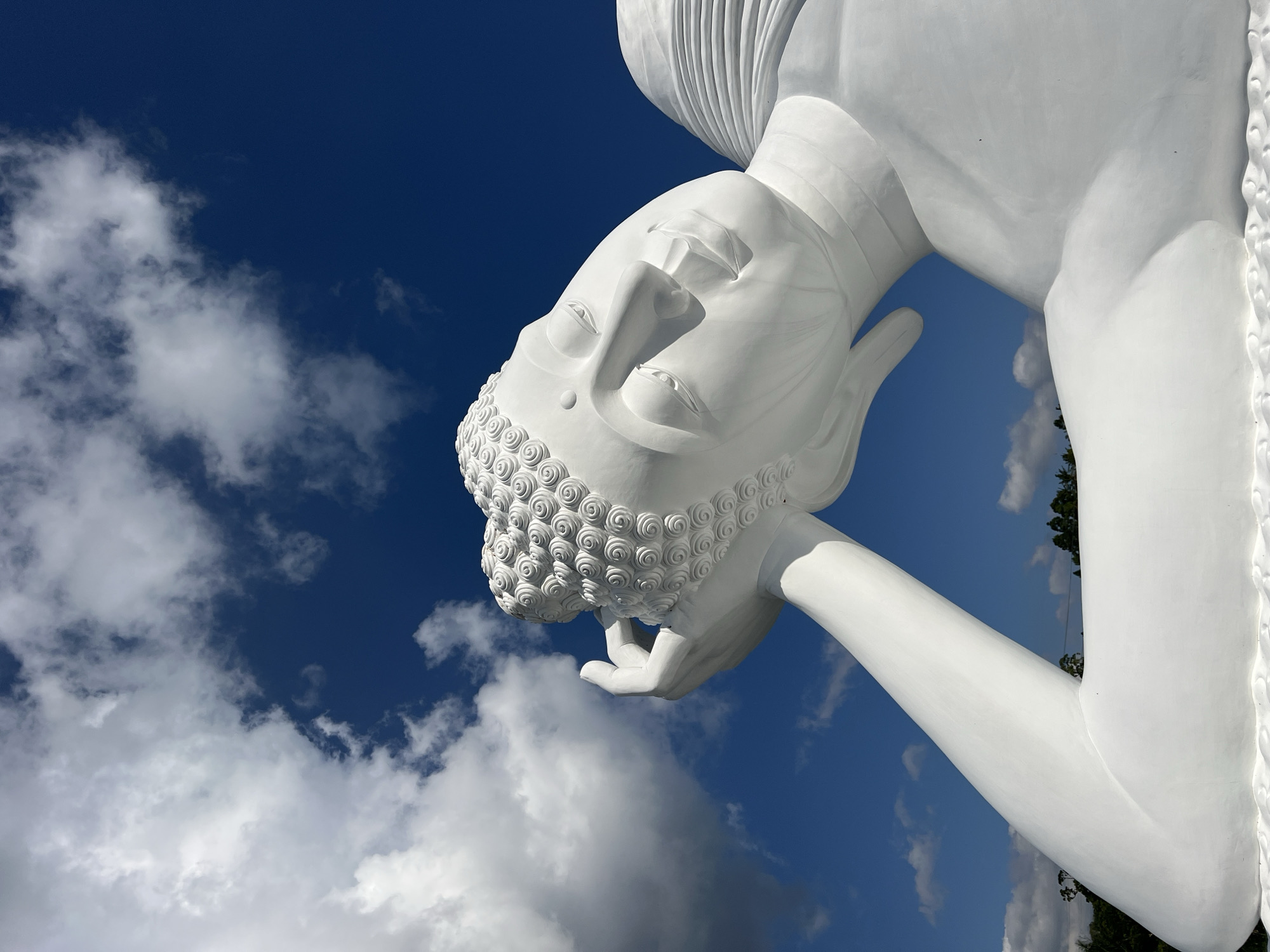 A low-angle, close-up shot captures the serene face of a colossal white reclining Buddha statue against a vibrant blue sky dotted with fluffy white clouds. The statue's head is tilted to the right, resting on its arm, with its eyes closed in peaceful contemplation. The intricate details of the Buddha's curls are visible on its head, and its facial features are finely sculpted. The statue appears to be located outdoors, likely in an open area, given the sky and glimpses of greenery and trees at the bottom edge of the image. The lighting suggests a sunny day, casting soft shadows on the statue's form. The overall impression is one of tranquility and grandeur. This statue is located in Pupuan, Indonesia.