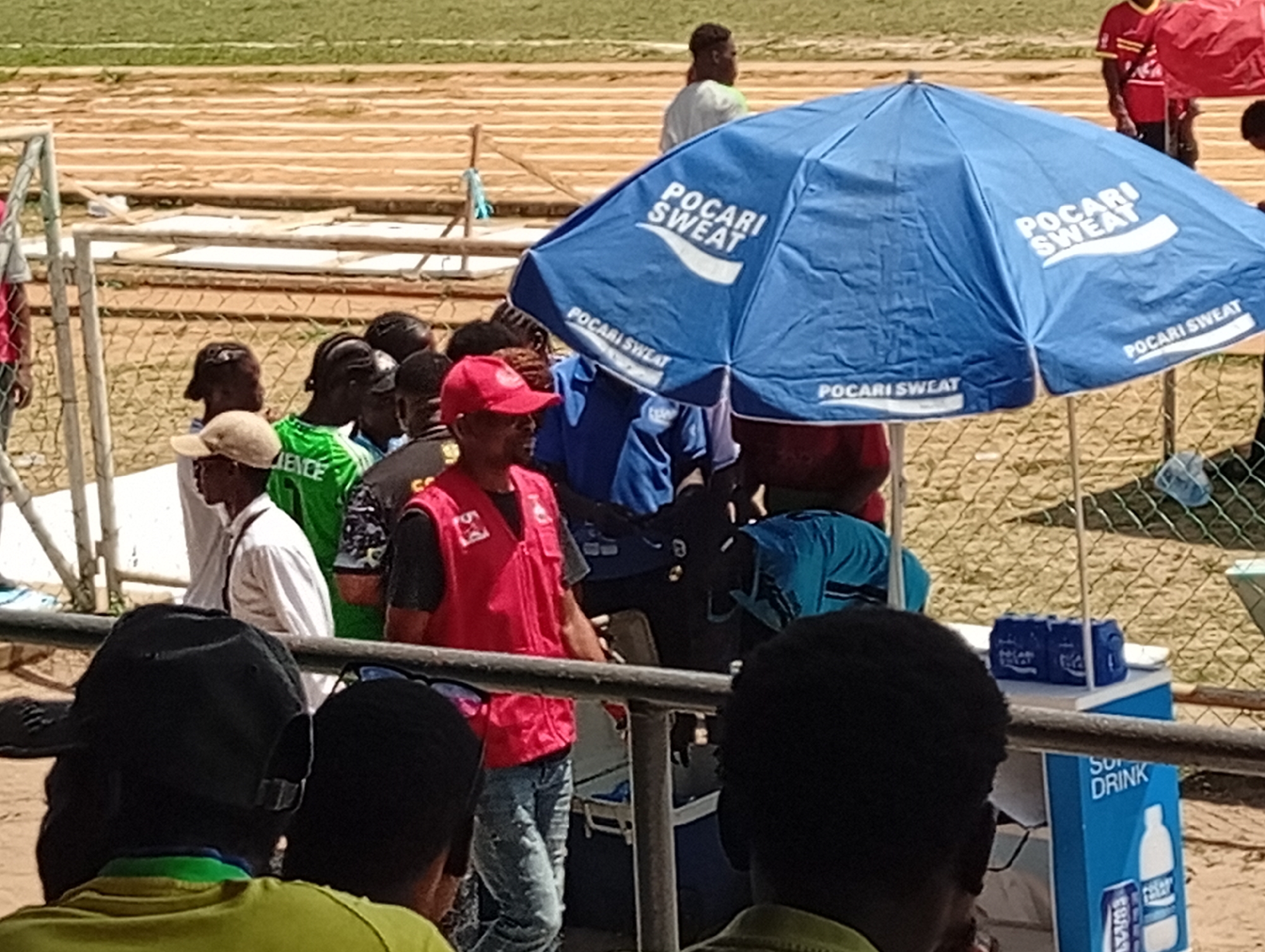 The image shows an outdoor scene at what appears to be a sporting event, possibly a track and field competition, with wooden bleachers in the background. The dominant feature is a blue umbrella with "POCARI SWEAT" branding, sheltering a refreshments stand. The stand has a blue cooler with "SUPER DRINK" printed on it and is stocked with drinks. Several people are visible in the foreground and middle ground. One man wears a red vest and a red cap. Others are wearing various casual clothes and caps. The scene suggests a hot day. Although specific details confirming the location as Lagos, Nigeria, are absent, the general environment and demographics are consistent with that location.