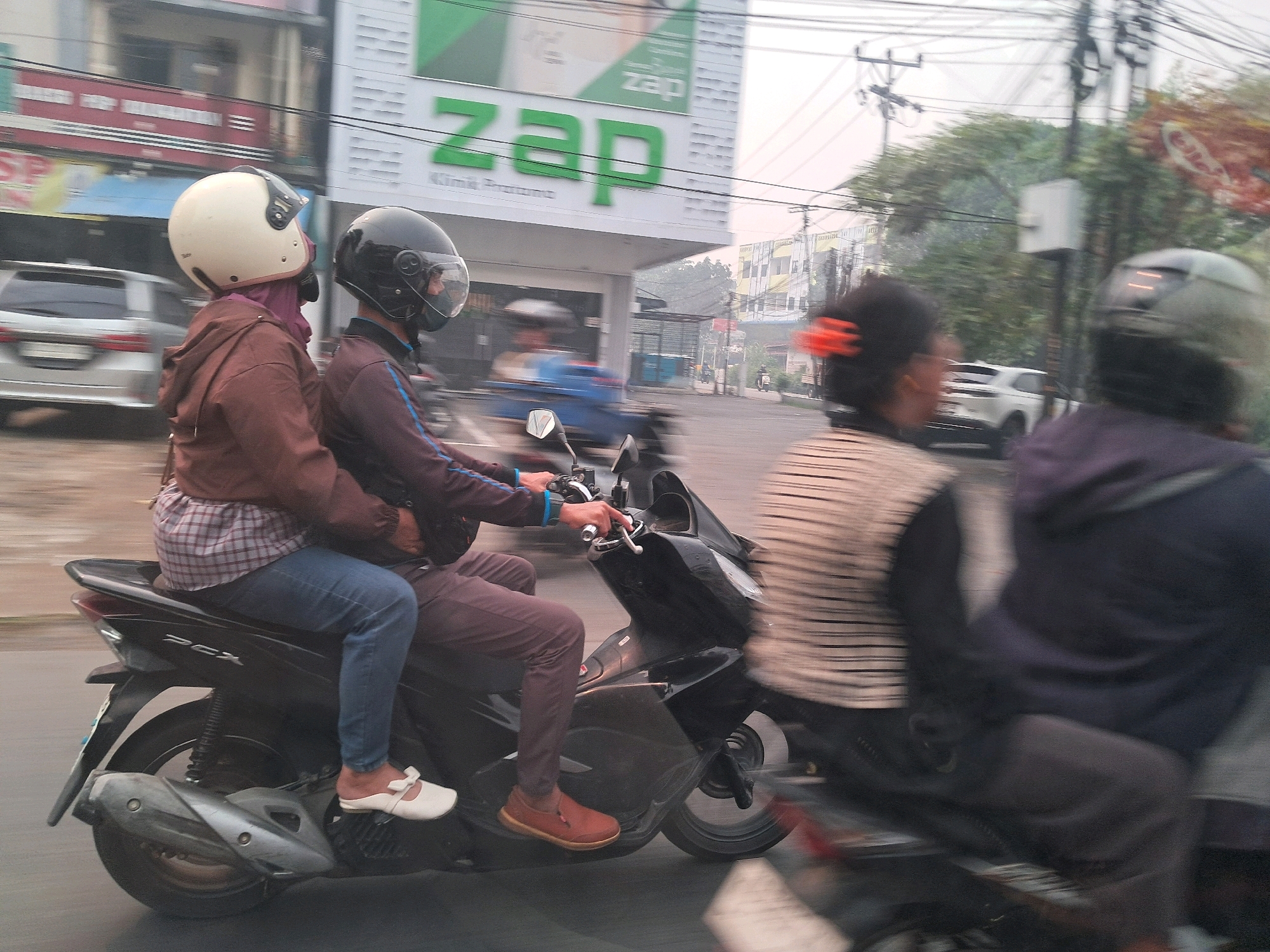 On a street in Pontianak, Indonesia, two people ride a black motorcycle. The rider in the front wears a black helmet and a dark jacket. The passenger behind them has a beige helmet and a brown jacket. The passenger's left leg is visible, wearing blue jeans and a white shoe. The motorcycle has the "PCX" logo visible. In the background, a silver car is parked, and a building with a green and white sign displaying the word "zap" in large green letters is visible. Traffic lights and power lines crisscross the sky. The scene is captured with motion blur, indicating the vehicles are in motion.