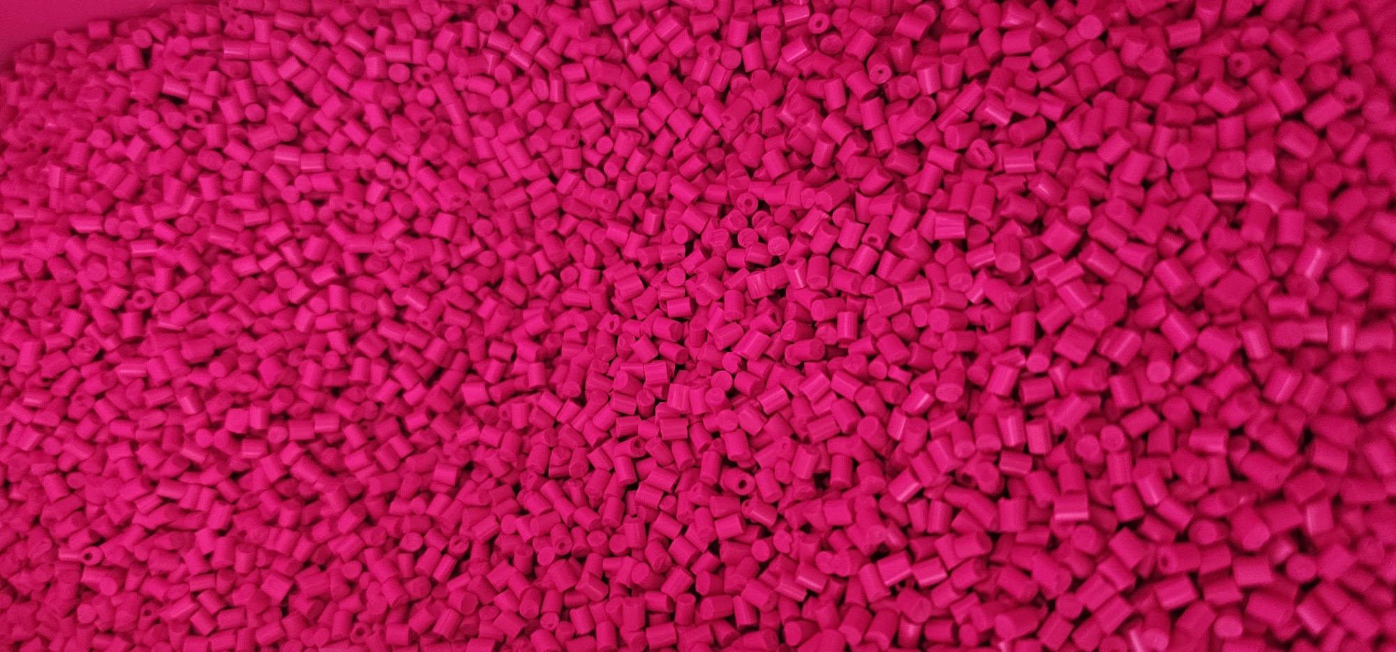 The image is a close-up, top-down view of a large pile of small, cylindrical, bright pink plastic pellets. The pellets are of uniform size and shape, with rounded ends, and are densely packed together, filling the entire frame. The lighting creates some shadowing and highlights, giving the pile a textured appearance. There are no people, animals, or any other objects visible in the image. The background is entirely composed of the same pink pellets. The image does not provide any information about its location in the city of Löhne, Germany, or any context beyond the visual content of the plastic pellets.