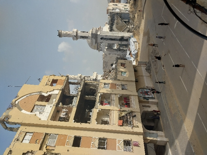 This image depicts a scene in Khan Yunis, Palestinian Territory, characterized by extensive urban destruction under a clear, light blue sky.
In the foreground and to the left, a multi-story, beige-colored building shows severe structural damage. Its upper floors are significantly compromised, with large sections of walls missing, exposing interior spaces and rebar. A distinctive arched opening is visible on one of the higher levels, revealing patterned interior walls. Several balconies on lower floors are partially intact, with some exhibiting hanging laundry. The ground floor features an arched entrance and a vibrant mural on a wall next to it. Piles of rubble are accumulated at the base of this building and along the adjacent dirt/gravel area.
In the mid-ground, to the right of the damaged building, stands a partially destroyed mosque or religious structure. Its prominent, white minaret remains largely intact, though visible damage is present on its upper section. The main body of the mosque shows significant destruction, with a large portion of its dome or roof collapsed, and debris scattered around its base.
The foreground features an asphalt street with white lane markings. Scattered across this street are approximately seven to eight individuals, primarily young males. One individual in the center foreground appears to be standing alone. Another group of three to four individuals is further down the street, seemingly engaged in an activity involving a small, dark object that resembles a ball. Near a pile of rubble on the right side of the street, one person is crouched down. Near the ground floor of the damaged building on the left, a smaller group of three to four people, including adults and possibly children, are visible near the entrance. The overall scene conveys a post-conflict environment with civilian activity amidst widespread structural devastation.