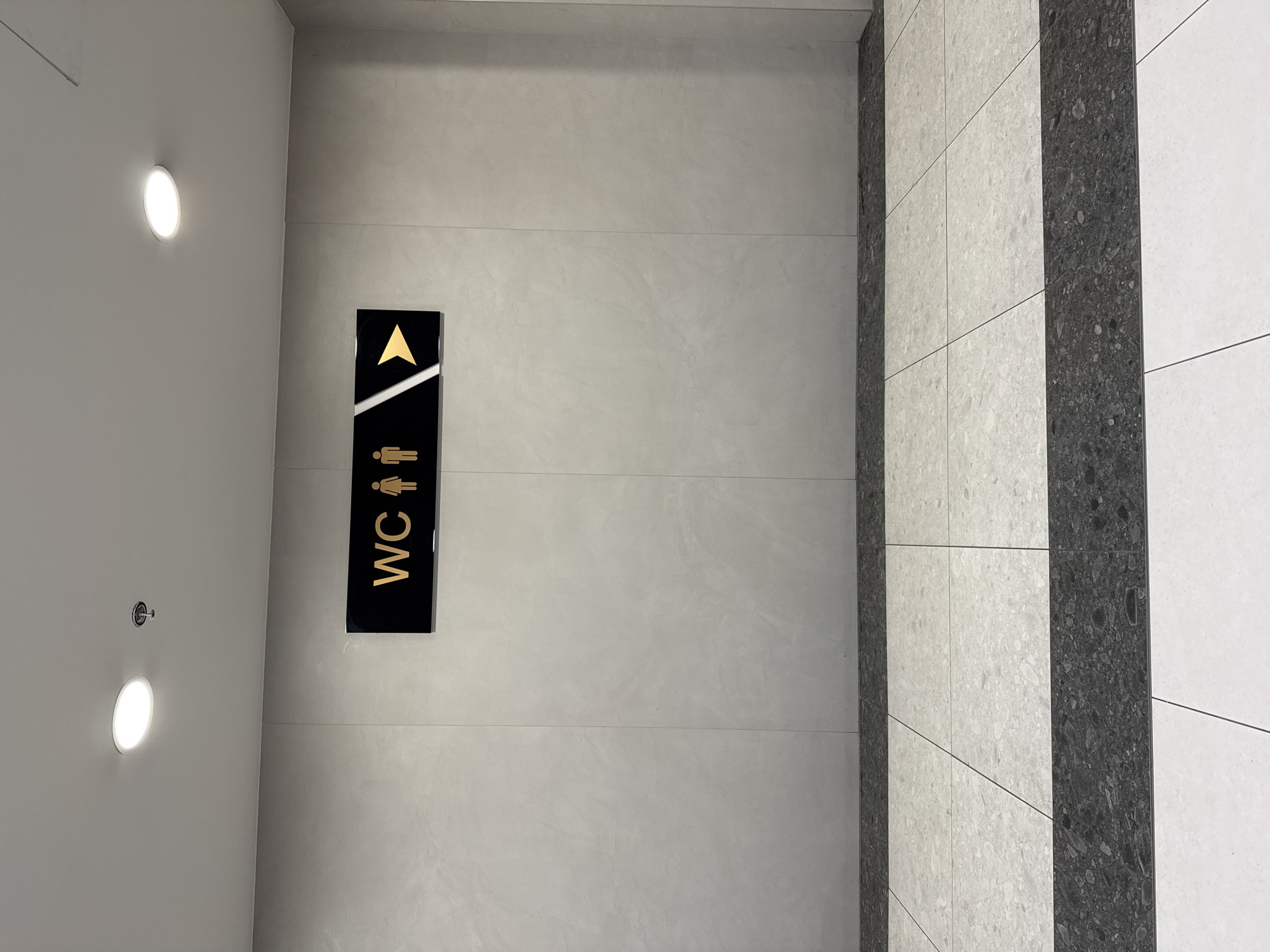 An indoor, well-lit scene depicts a modern hallway or corridor, likely within a public building in Burgas, Bulgaria. The focal point is a black rectangular sign mounted vertically on a light grey wall. The sign, rendered in gold, features an upward-pointing arrow at the top, followed by a gender-neutral symbol depicting combined male and female figures. Below these, the letters "WC" are stacked vertically. A white diagonal stripe adds a contemporary design element to the sign's upper section.
The walls are composed of large, smooth light grey panels, while the ceiling, also light grey, features two illuminated circular recessed lights. A small sprinkler head is also visible on the ceiling. The floor presents a striking pattern of rectangular tiles: a wide central strip of dark grey, heavily speckled tiles is flanked by lighter grey or white speckled tiles, which then transition to plain white tiles on the far right. No people are visible, and the scene implies a directional path to a restroom ("WC"). The overall aesthetic is clean, modern, and functional.