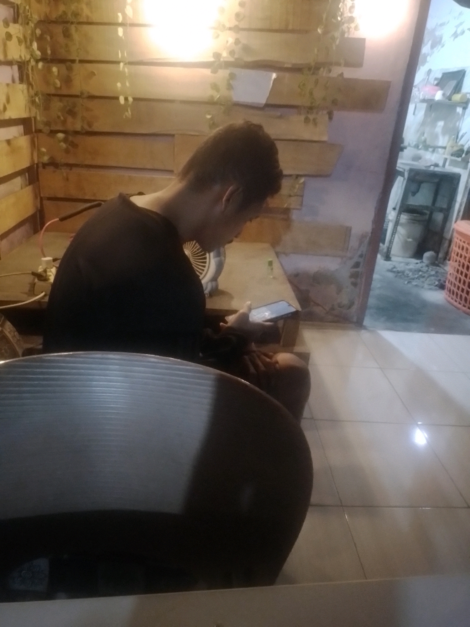 A person, viewed from the side and from behind, is seated and looking at their phone. They are wearing a black long-sleeved shirt. The background consists of wooden slat walls with decorative greenery and a bright light source above. To the right, an open doorway reveals a glimpse of another room with shelves, a bucket, and rubble on the floor. In the foreground, a portion of a metallic chair is visible. The floor is tiled.