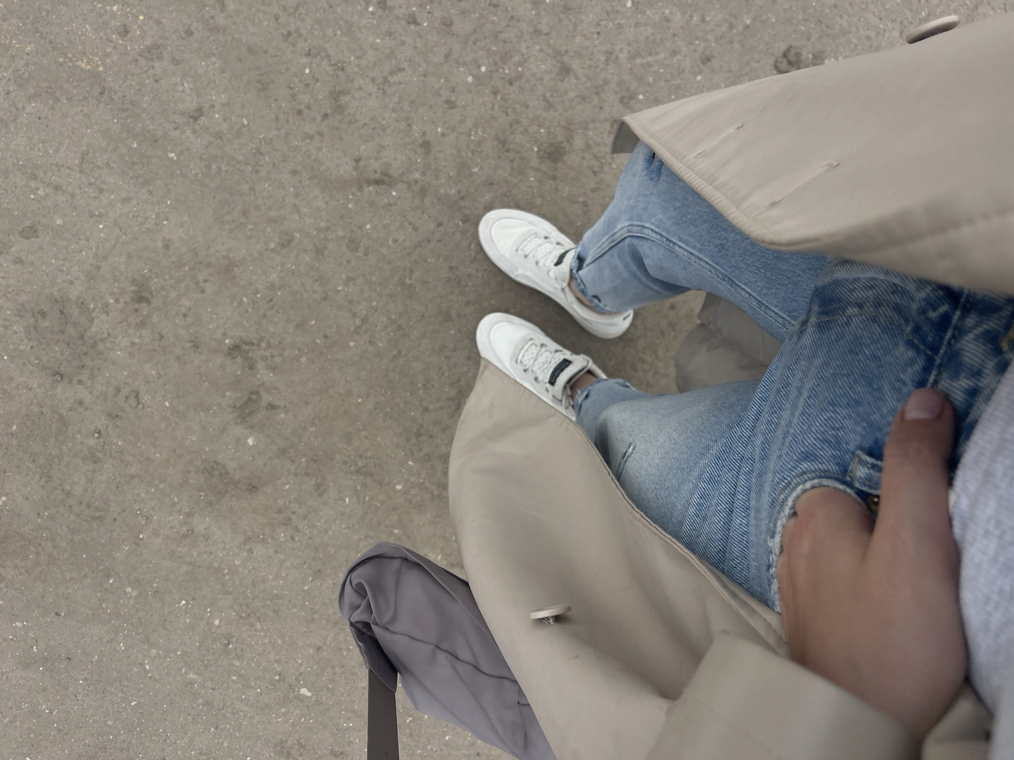 The image presents a top-down view of a person seated on a likely urban surface, wearing light blue jeans and white sneakers. A light beige trench coat is draped over the legs. The individual's hand is partially visible near the jeans pocket. A dark grey tote bag rests adjacent to the trench coat. The setting is a paved area, with no discernible indicators that would link it to Molokovo, Russia. No actions or interactions can be observed in the image.