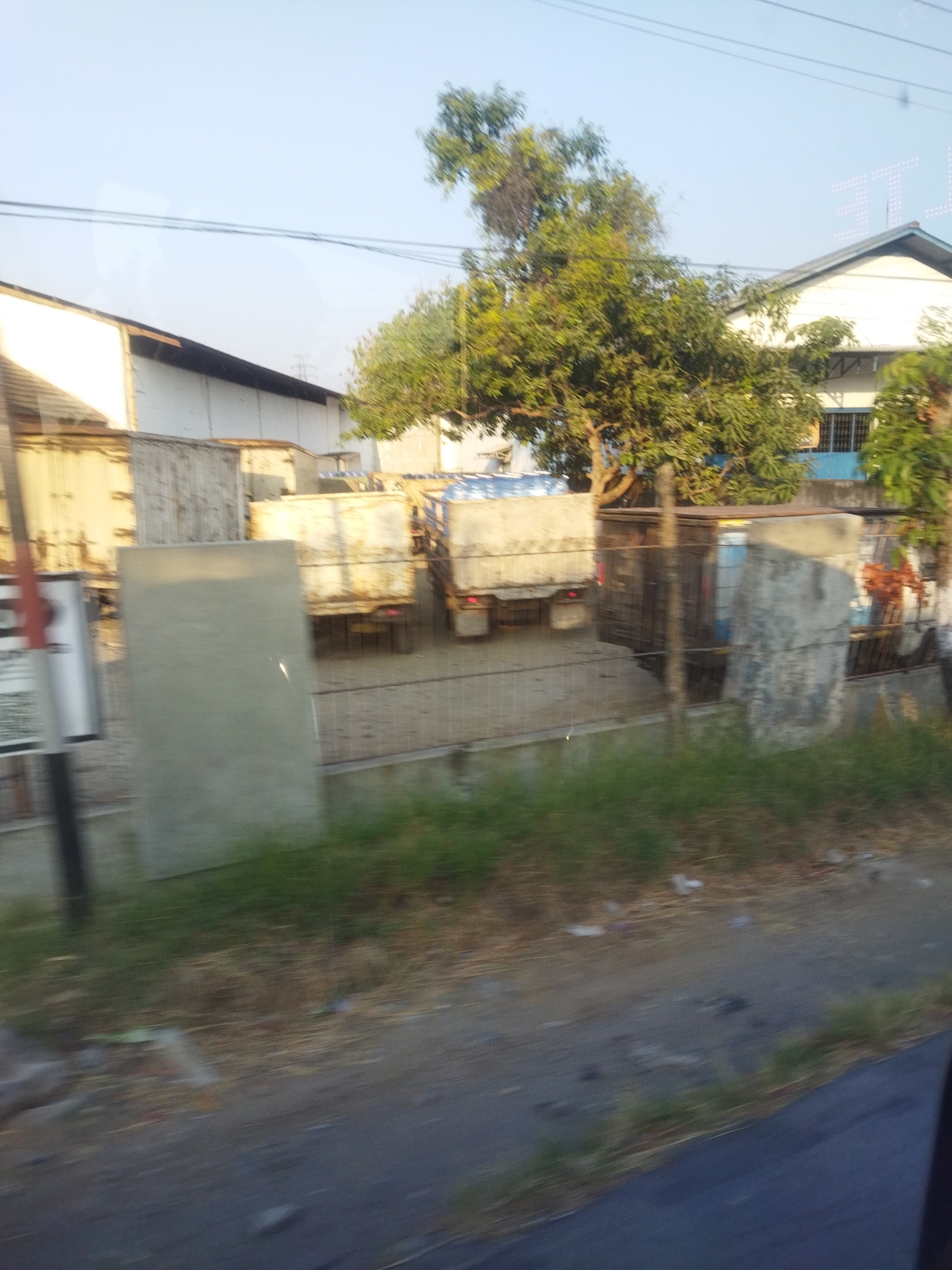 The image shows an outdoor scene in Selogiri, Indonesia, with several cargo trucks parked in a yard. The trucks appear to be carrying goods, with some containers partially visible. In the background, a building with a white facade and a dark roof is present. Trees with green foliage are scattered around the area, suggesting a semi-urban or industrial setting. The ground is a mixture of dirt and some sparse vegetation. The sky is clear and pale blue, indicating daylight.