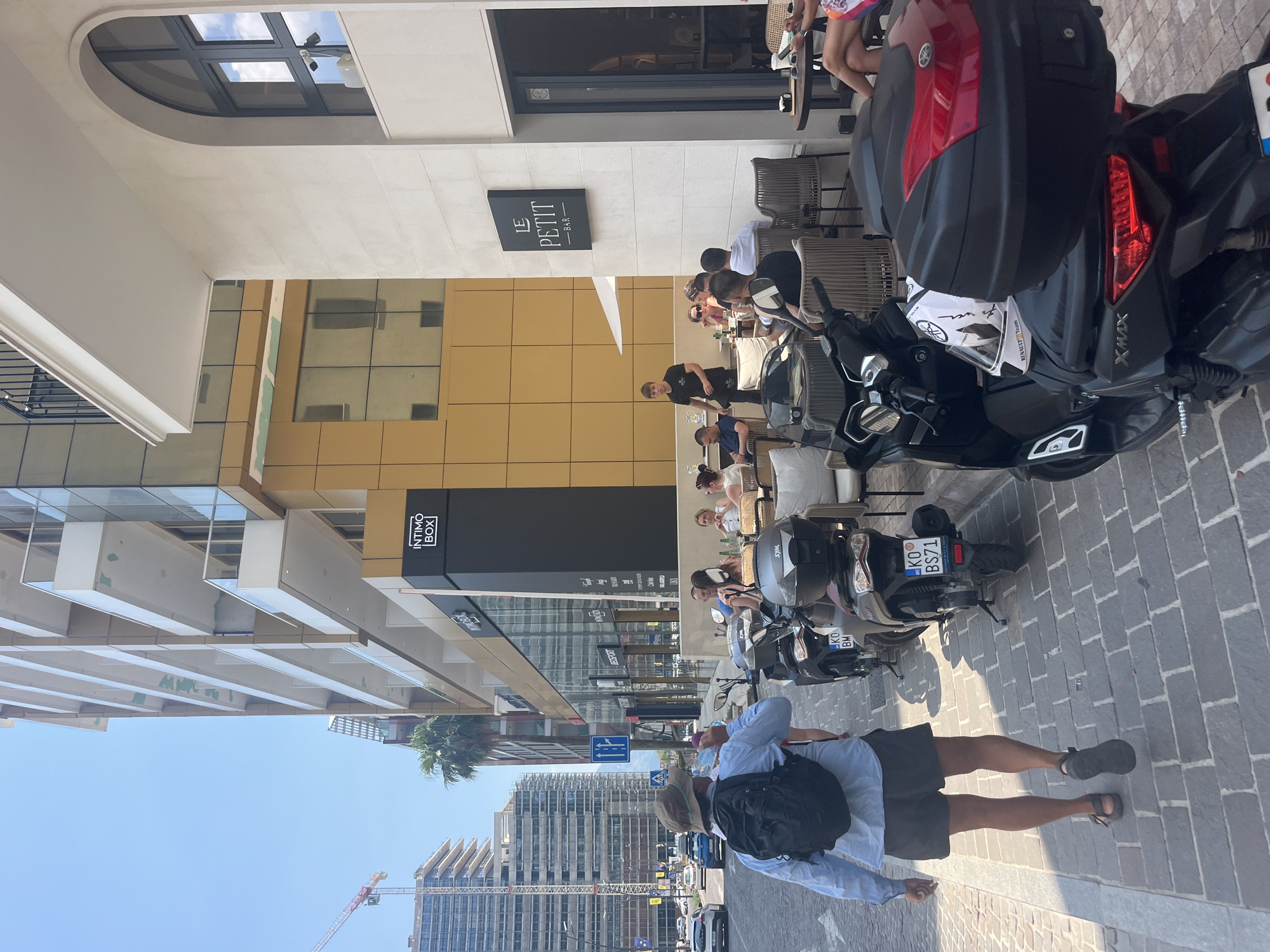 The image depicts an outdoor scene in what appears to be a city, possibly during daytime given the bright sunlight casting shadows. In the foreground, a person is walking away from the camera, wearing a light blue shirt, dark shorts, a hat, and a backpack. The person is walking on a paved sidewalk made of grey cobblestones.
Behind the person, several scooters are parked. One scooter, a Yamaha XMAX, is prominently featured, with a red and black top box. It has a license plate that reads "KO BS71". Another scooter is partially visible behind it.
The background reveals a street scene with buildings and people. A building with a modern facade, featuring large glass windows and cream-colored panels, dominates the left side of the image. A sign that reads "LE PETIT BAR" is visible on a white tiled wall. Further down the street, another building with a dark facade has a sign that says "INTIMO BOX". People are seated at tables outside what appears to be a cafe or bar, suggesting a leisurely or social atmosphere.
In the distance, construction is underway on tall buildings, indicated by a crane. The sky is clear and blue, suggesting fair weather. The overall impression is of a bustling urban environment on a pleasant day.