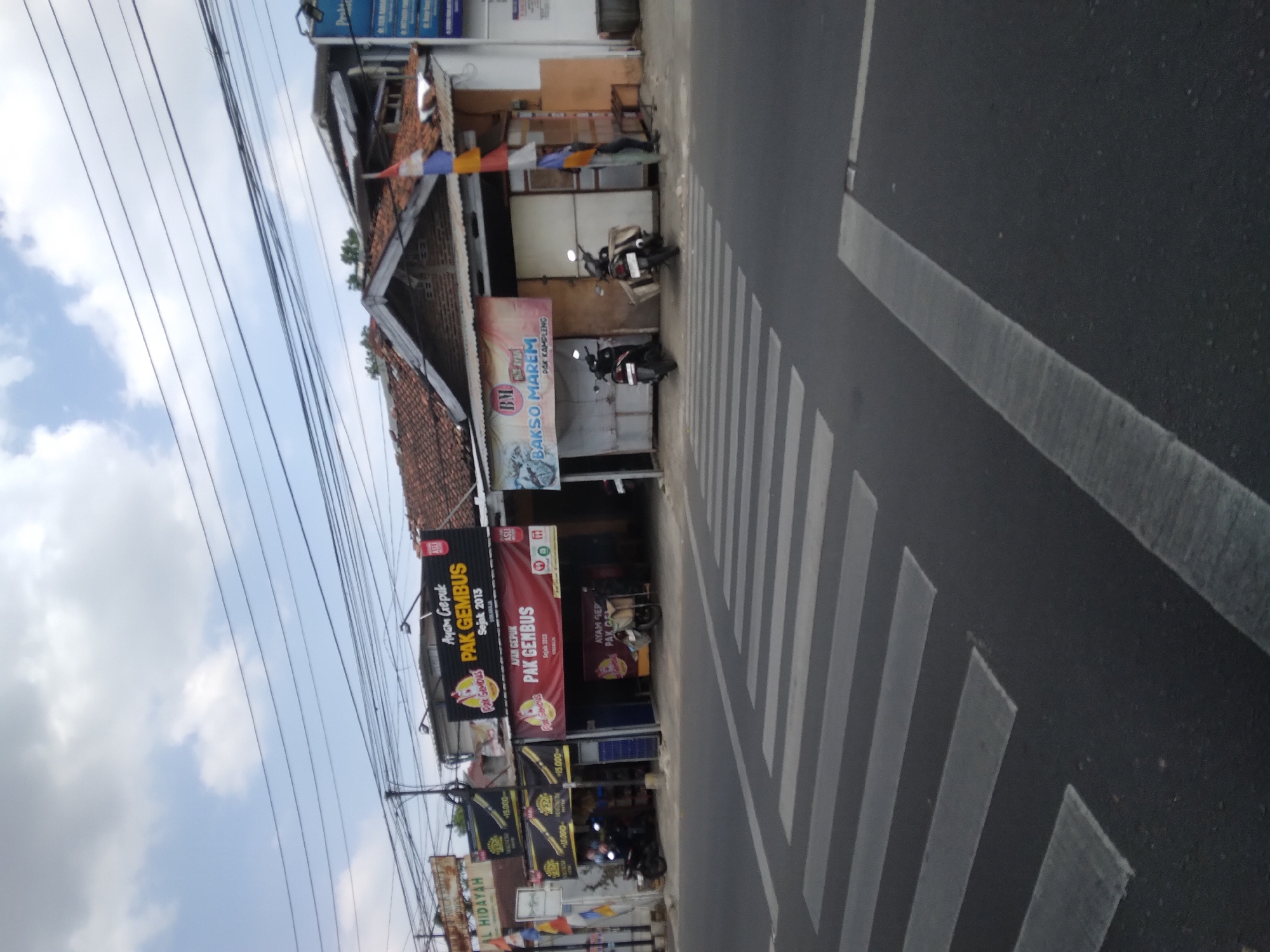 The image shows a street scene in Berkoh, Indonesia. The street has a pedestrian crossing in the foreground. On the right side of the street, there are several businesses with awnings and signage. One sign reads "AYAM GEPUK PAK GEMBUS" which indicates a restaurant selling fried chicken. Another sign says "BAKSO MAREM PAK KAMLENG", suggesting a place selling meatballs. Several motorcycles are parked in front of the establishments. The sky is partly cloudy.