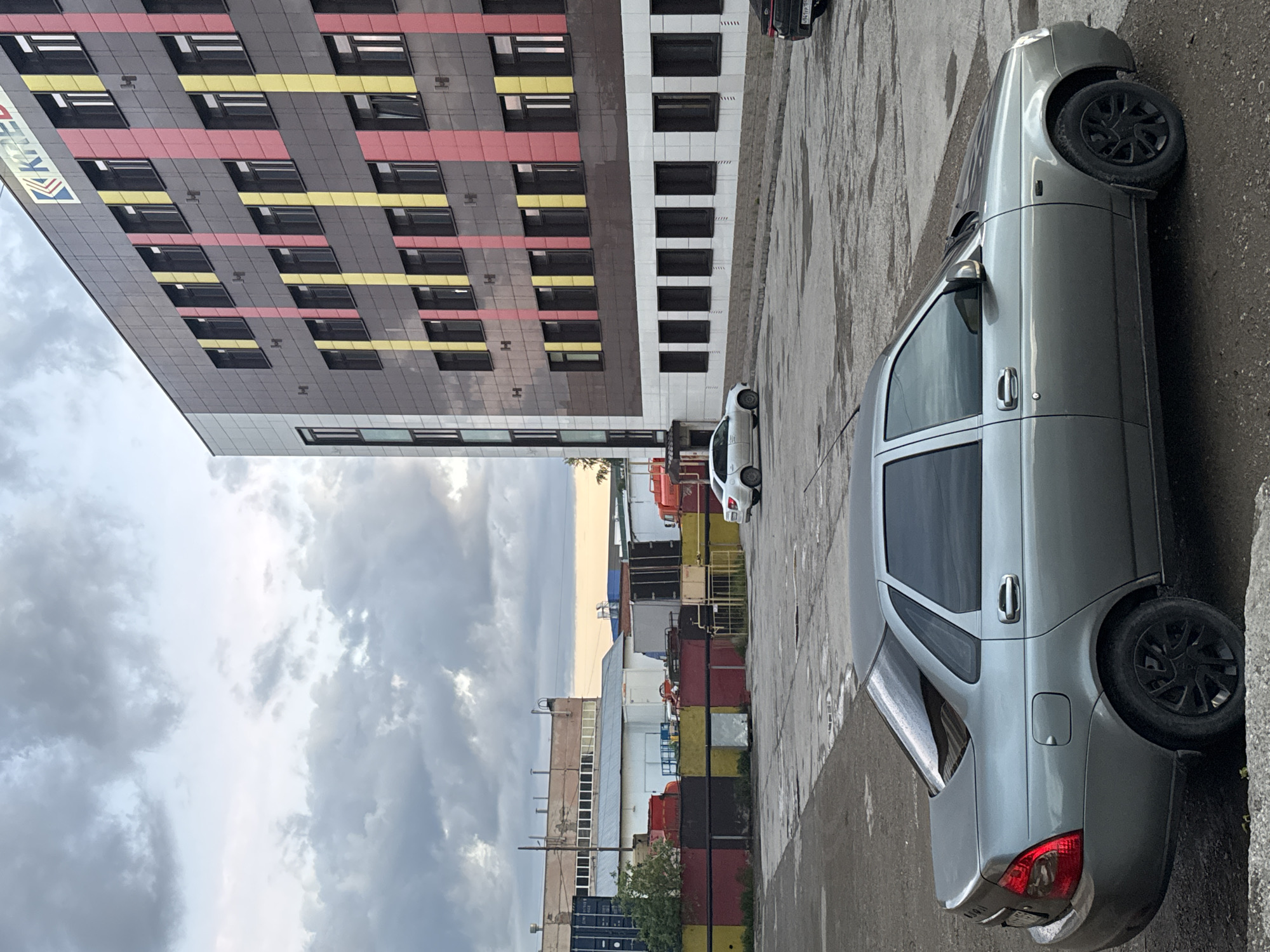 The image displays an outdoor scene in what appears to be an urban or industrial area, consistent with Perm, Russia, under an overcast sky filled with grey clouds.
In the foreground and to the right, a light grey sedan, possibly a Lada, is parked on a paved surface that looks wet or stained. The car features black multi-spoke rims, and its rear bumper and taillight are visible. Another white car is partially seen parked further back, while a dark vehicle with an unreadable license plate is partially visible at the far right edge of the frame.
To the left, a modern multi-story building stands prominently. Its facade is composed of dark grey panels accented by vibrant horizontal red and yellow stripes, with numerous rectangular windows interspersed across its surface. The word "КРЕА" (KREA) is distinctly visible on the upper left side of this building. Adjacent to it on the right is another building, presenting a white facade with a grid of dark, rectangular windows.
The middle ground reveals an industrial or commercial zone characterized by various shipping containers stacked in different colors, including red, yellow, and black. Additional industrial structures and buildings are also visible, suggesting a logistical or manufacturing environment.
The scene depicts a cloudy daytime setting, with no people visible and the primary activity being the parking of vehicles.
