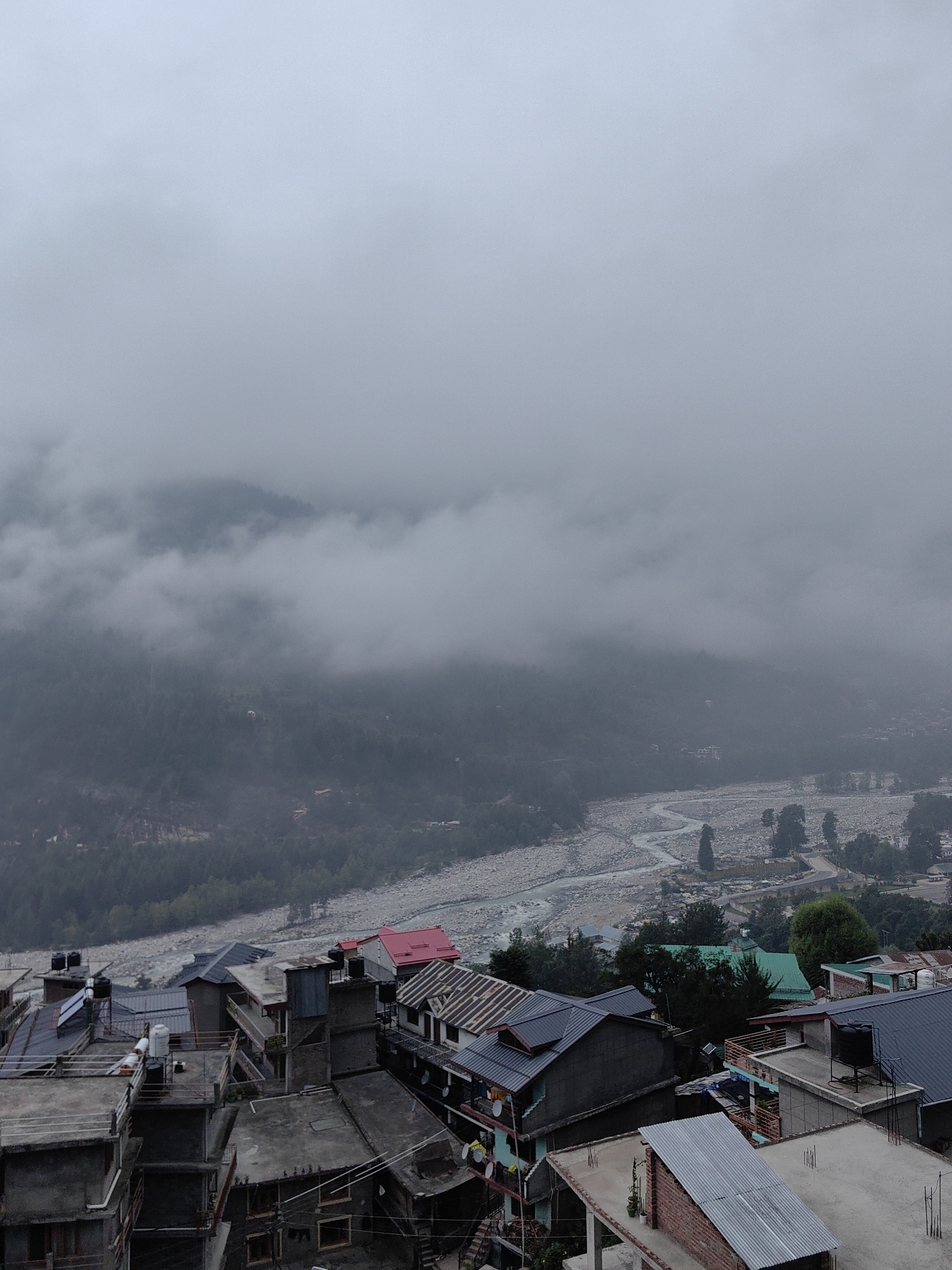 This is an elevated, wide-angle, outdoor, full-shot photograph captured in Vashisht, India, on an overcast day. The scene depicts a hillside town with dense cloud cover obscuring the upper portions of the surrounding mountains. In the foreground, the roofs of numerous buildings, varying in architectural style and material, are visible. Many of these buildings have visible water tanks on their rooftops. A river, appearing wide and stony, flows through the valley below. The town is situated on a steep incline, with the river meandering through the valley floor and a dense forest on the opposite bank. The sky is completely covered in thick, gray clouds, contributing to the muted, atmospheric light of the scene. There are no visible people or animals.