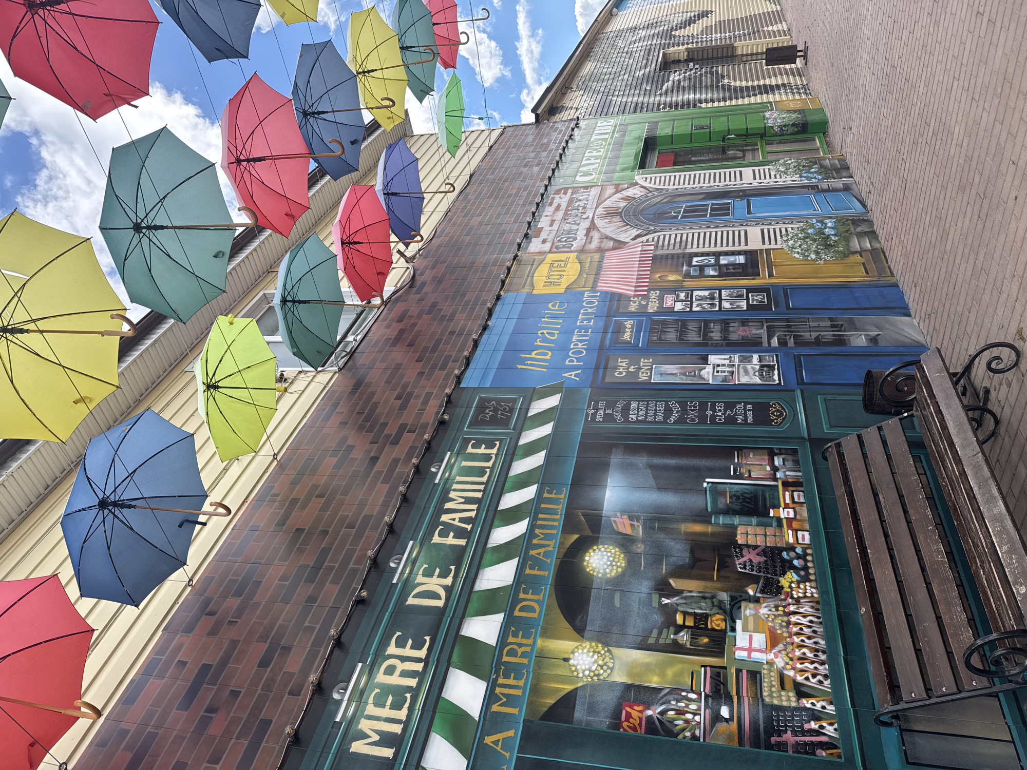 The image depicts a street scene with an artistic display of colorful umbrellas suspended above a painted facade that mimics storefronts. The facade features inscriptions in French, including "MERE DE FAMILLE" and "Librairie A PORTE ETROIT." The faux storefront windows showcase painted depictions of pastries and books. A bench sits in front of the facade, suggesting an invitation for passersby to pause and enjoy the display. The street appears to be made of brick, and the sky above is partly cloudy. The location is Osa, Russia.