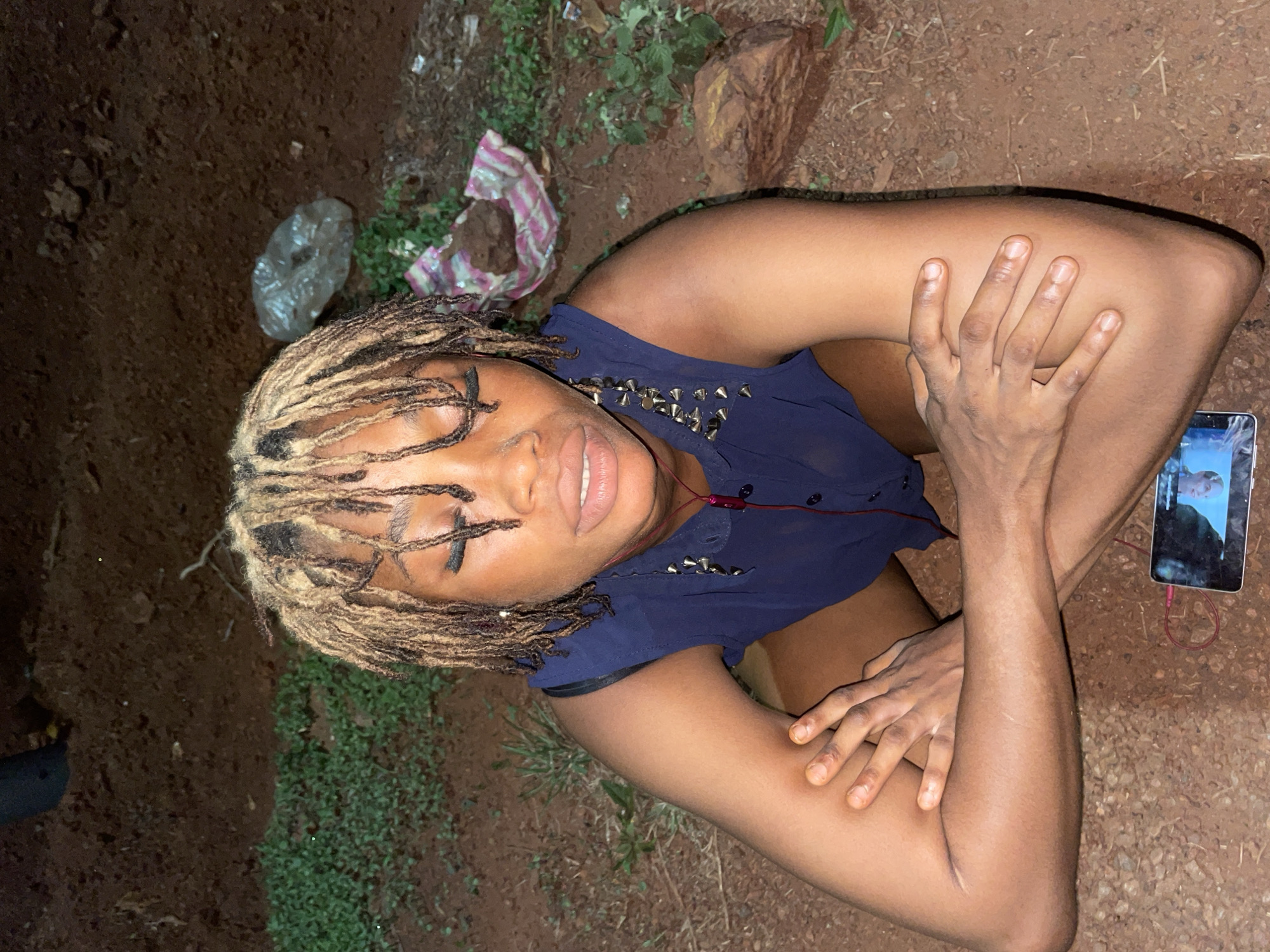 A medium shot depicts a person with tan skin, blonde dreadlocks, and a blue shirt with silver studs, lying on the ground in an outdoor location identified as Ogbe, Nigeria. The person's eyes are closed, and they are smiling slightly with closed lips. Their arms are crossed and resting on their knees. A smartphone connected to red earphones is located on the ground near their right arm. The ground consists of dark soil, scattered with small patches of green plants and debris, including a transparent plastic bag and striped plastic wrap. The angle of the image suggests the camera was positioned directly above the subject.