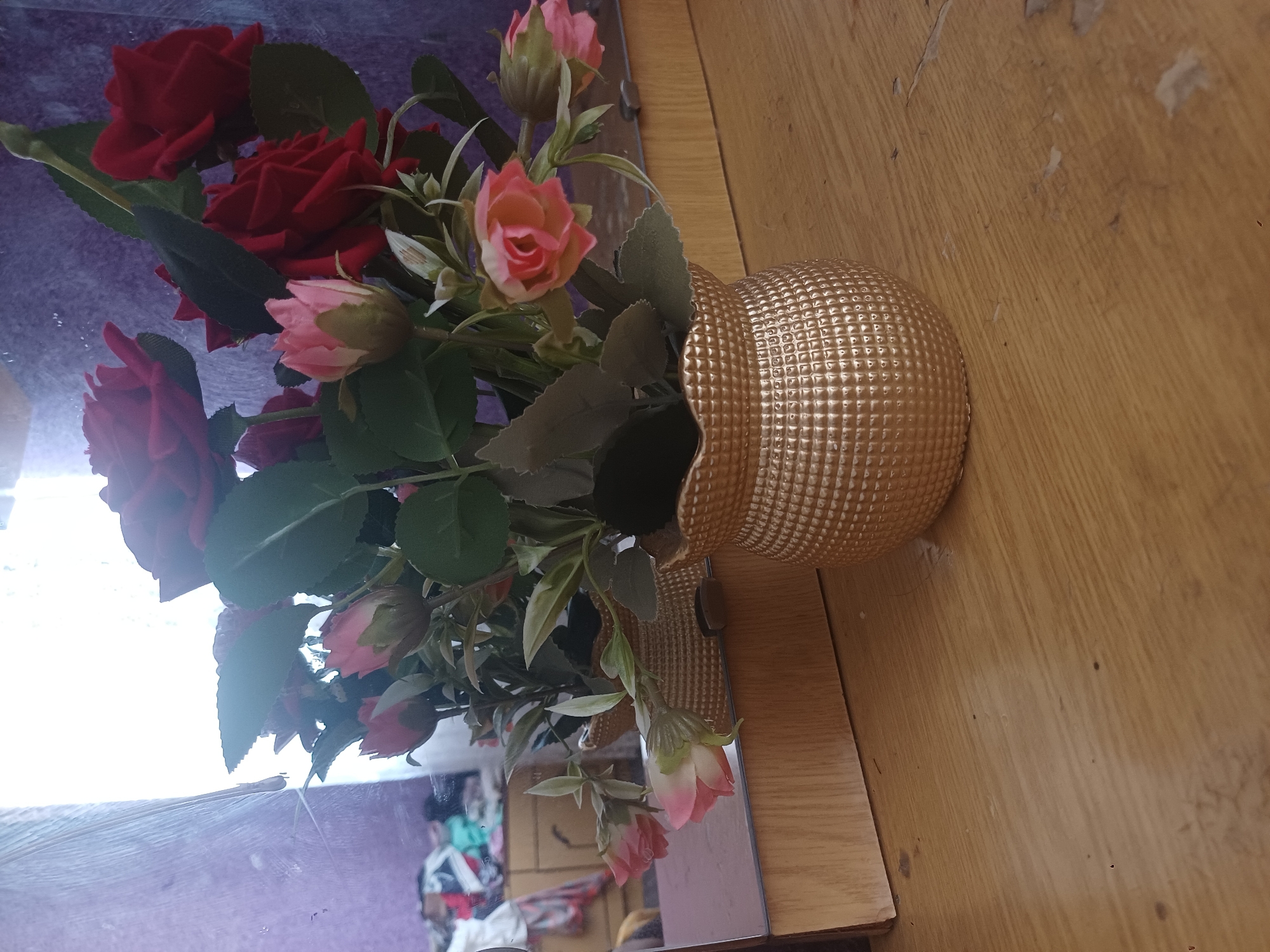 The image shows a bouquet of artificial red and pink roses in a textured golden vase. The vase sits on a wooden surface, likely a table or dresser. Behind the vase, a mirror reflects the bouquet, and in the reflection, a blurry background shows what appears to be a wooden dresser with clothing draped over it. The overall setting suggests an indoor space. There is no information available that can locate this scene in Jumashuy, Uzbekistan.