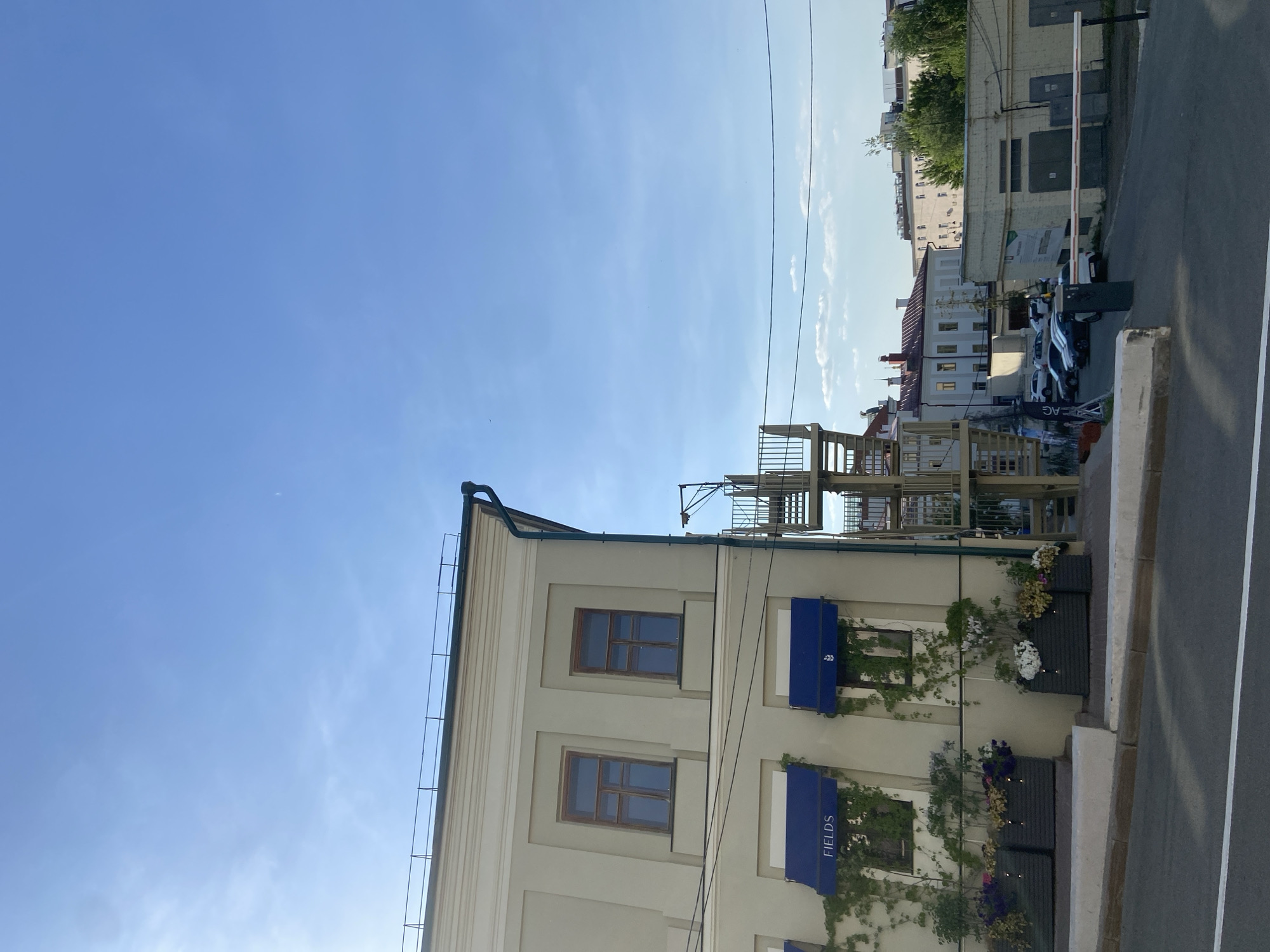 The image shows a multi-story building in Kazan, Russia. The building is painted in light beige and has multiple windows with brown frames. Blue awnings are installed above some of the windows, bearing the word "FIELDS". Green vines are growing around the awnings. A metal fire escape staircase is visible behind the building. Adjacent to the building is a road with a curb. Further in the background, other buildings can be seen, including some with red roofs. A parking area with several cars is also visible. The sky is blue with a few clouds. Overhead power lines are present in the sky.