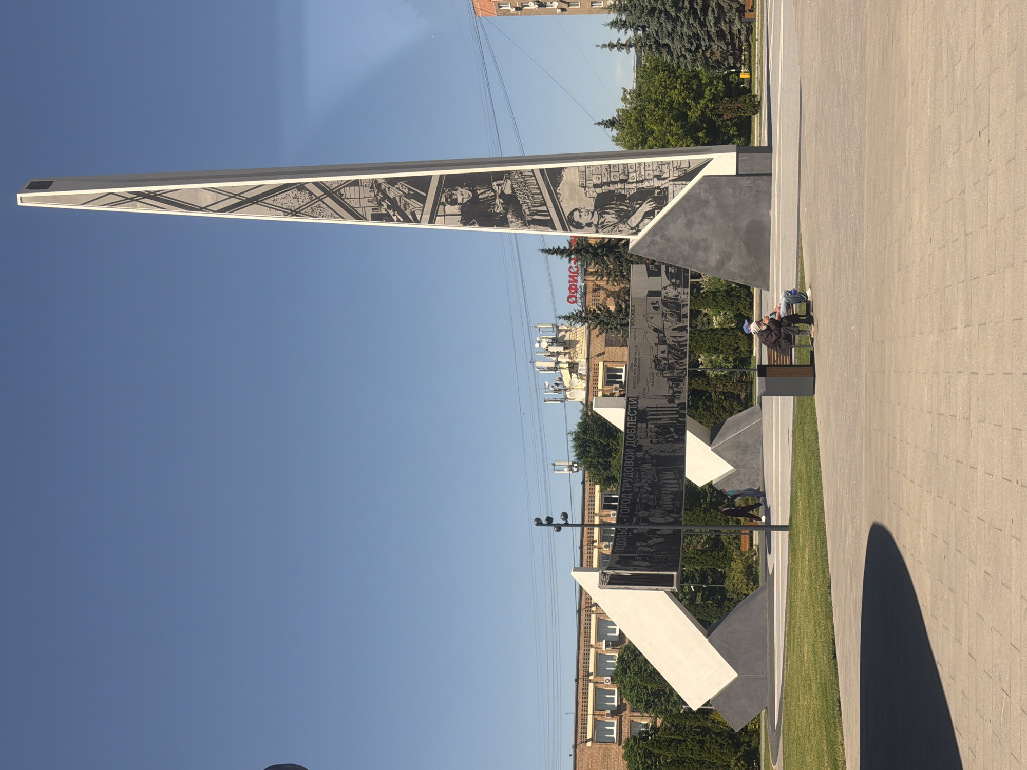 This image shows the "Podolsk - City of Labor Valor" monument, located in Podolsk, Russia. The monument is a tall, obelisk-like structure with relief carvings depicting scenes of labor and wartime effort. In front of the monument, a long, horizontal panel also features relief carvings. To the right of the main obelisk, two people are seated on a bench, facing away from the camera and towards the monument. One person is wearing a blue cap, and the other is wearing a dark hat. The environment is an outdoor urban setting with a clear blue sky, paved walkways, a grassy area, and trees. In the background, there are buildings and power lines.