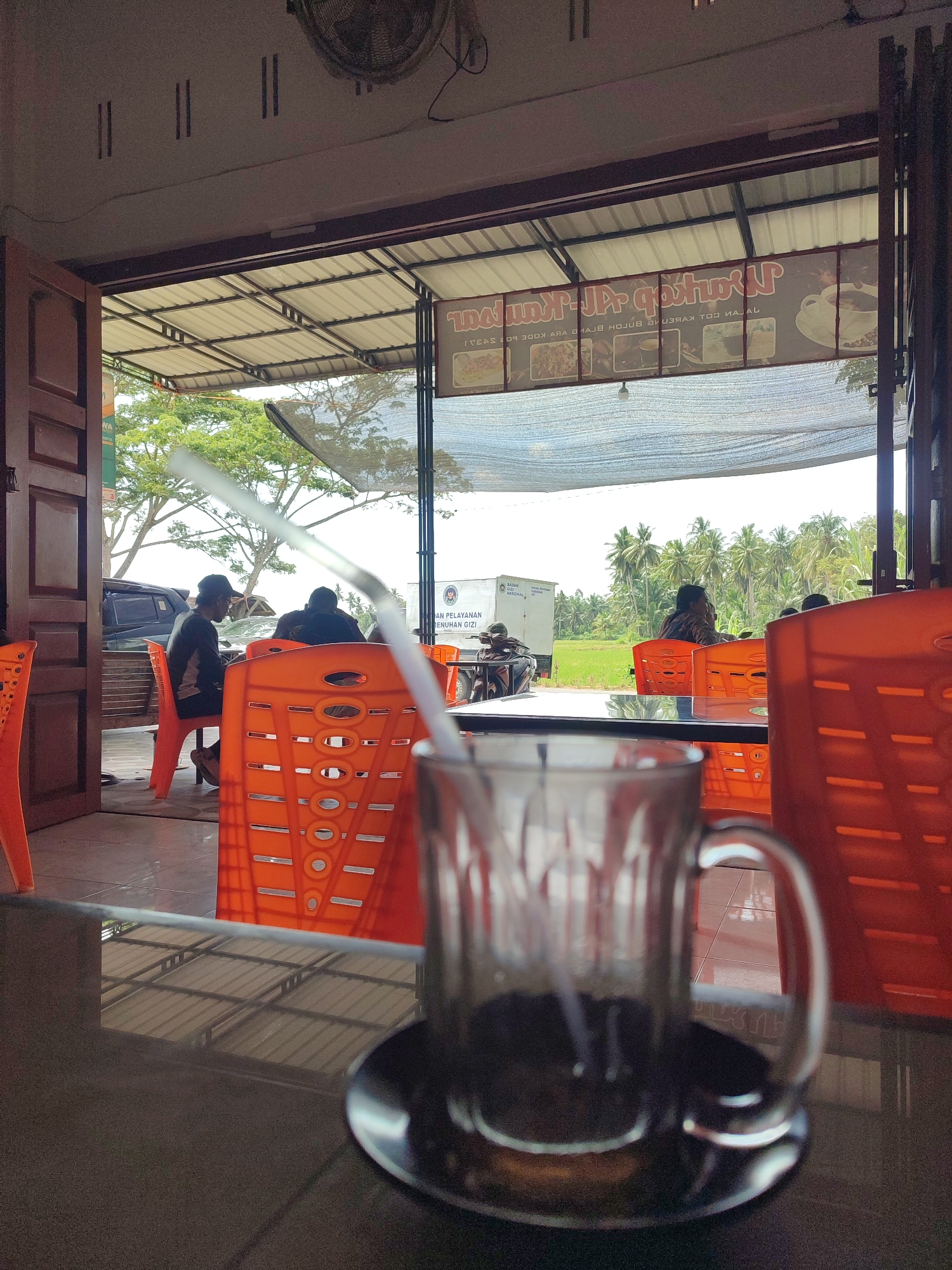 This image depicts a casual, open-air eatery or cafe in Kruengmanyang, Indonesia, observed from an indoor perspective. In the blurry foreground, a glass mug containing a dark liquid and a white straw rests on a dark table.
The midground features several vibrant orange plastic chairs, some occupied by patrons. People are seen relaxing in these chairs, facing outwards towards the bright exterior. One person on the left, wearing a dark shirt and cap, is partially visible, while another with a patterned shirt sits further back.
The background reveals a bright, sunny daytime scene with a lush tropical landscape. Numerous green trees, including a prominent cluster of palm trees, line the horizon. A green field, possibly a rice paddy, is visible before the distant trees. A white truck, bearing the partially visible text "AN PELAYANAN," "MENUHAN GIZI," and "NASIONAL," suggests a public service vehicle. A motorcycle is also partially visible near the truck.
The establishment has an open design with a light-colored corrugated metal roof supported by a dark frame, and a concrete floor. A ceiling fan is partially visible in the top left. Mirrored text from an overhead banner, reading "zostwa-JPqötu" and "1810930ОХ АЯА МАВ HOAX TOJ KAJAL," is reflected on the table surface. The clear, bright sky indicates a sunny day, consistent with a warm climate. The overall mood appears relaxed.