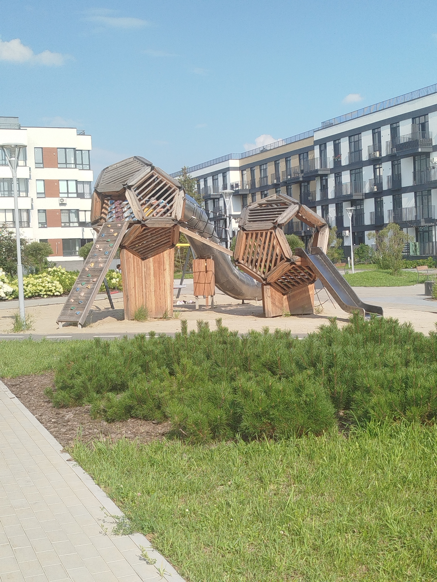 The image captures an outdoor contemporary playground area in the city of Tyumen, Russia. The playground features a modern, angular wooden climbing structure with an integrated slide and a climbing wall. The structure is set on a sandy surface, bordered by a paved walkway and a lush green lawn with low-lying pine shrubs. In the background, two modern multi-story apartment buildings stand prominently, one white with brown accents and the other white and gray with black detailing, their balconies and windows visible under a clear blue sky with scattered clouds. The scene appears to be a well-maintained residential or public space, devoid of any visible people or ongoing activities.