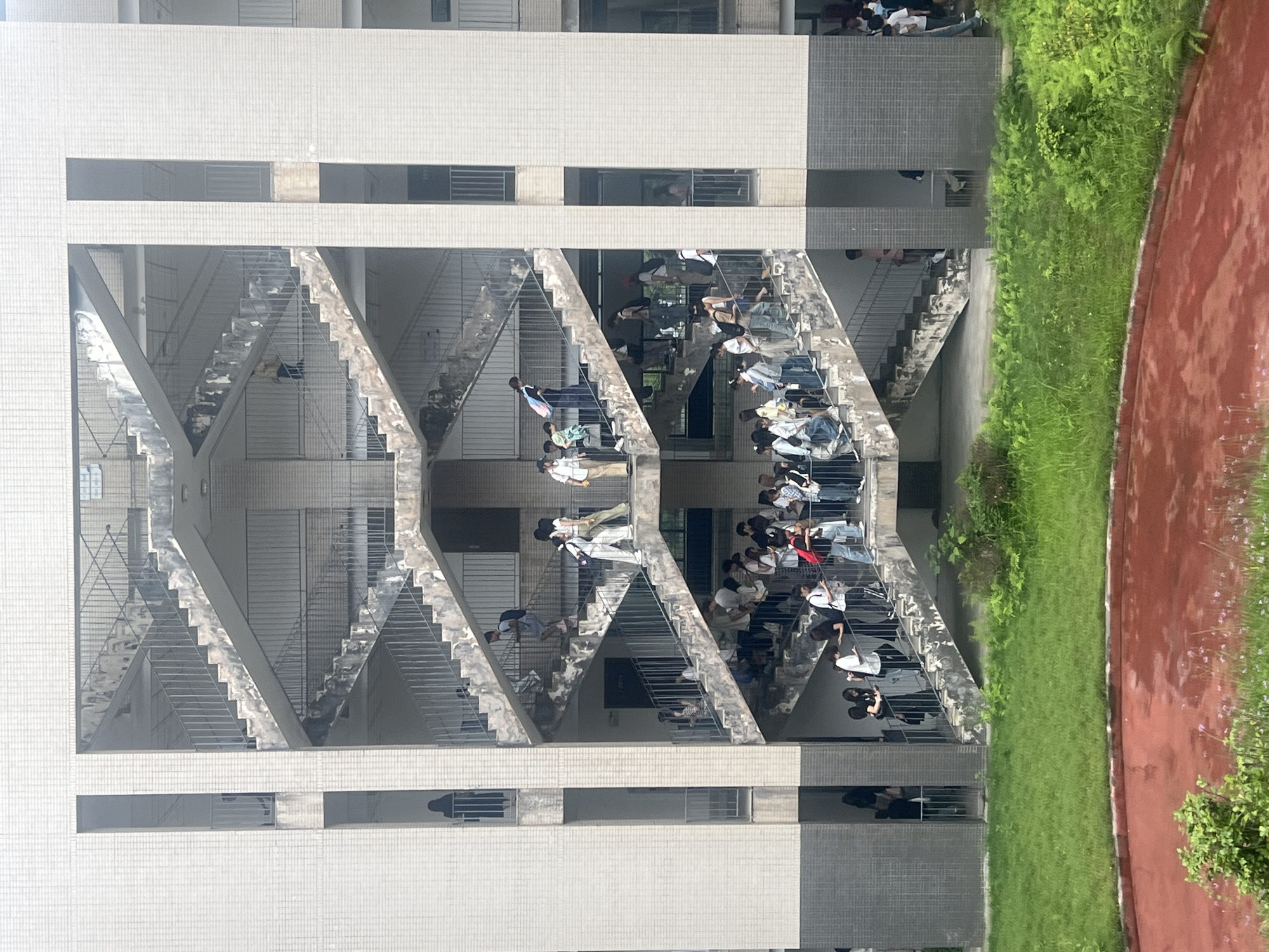 The image depicts the exterior of a multi-story building, likely a school, in Jiangjin, China, characterized by open-air staircases with metal railings. A large number of students are present on the stairs, with some appearing to be moving between levels. The architecture is predominantly concrete with tiled or textured exterior walls. The environment includes some greenery and a running track adjacent to the building. Windows are visible on the building's facade, and some individuals are observable looking out. The scene suggests a period of transition, such as a class change or break time.