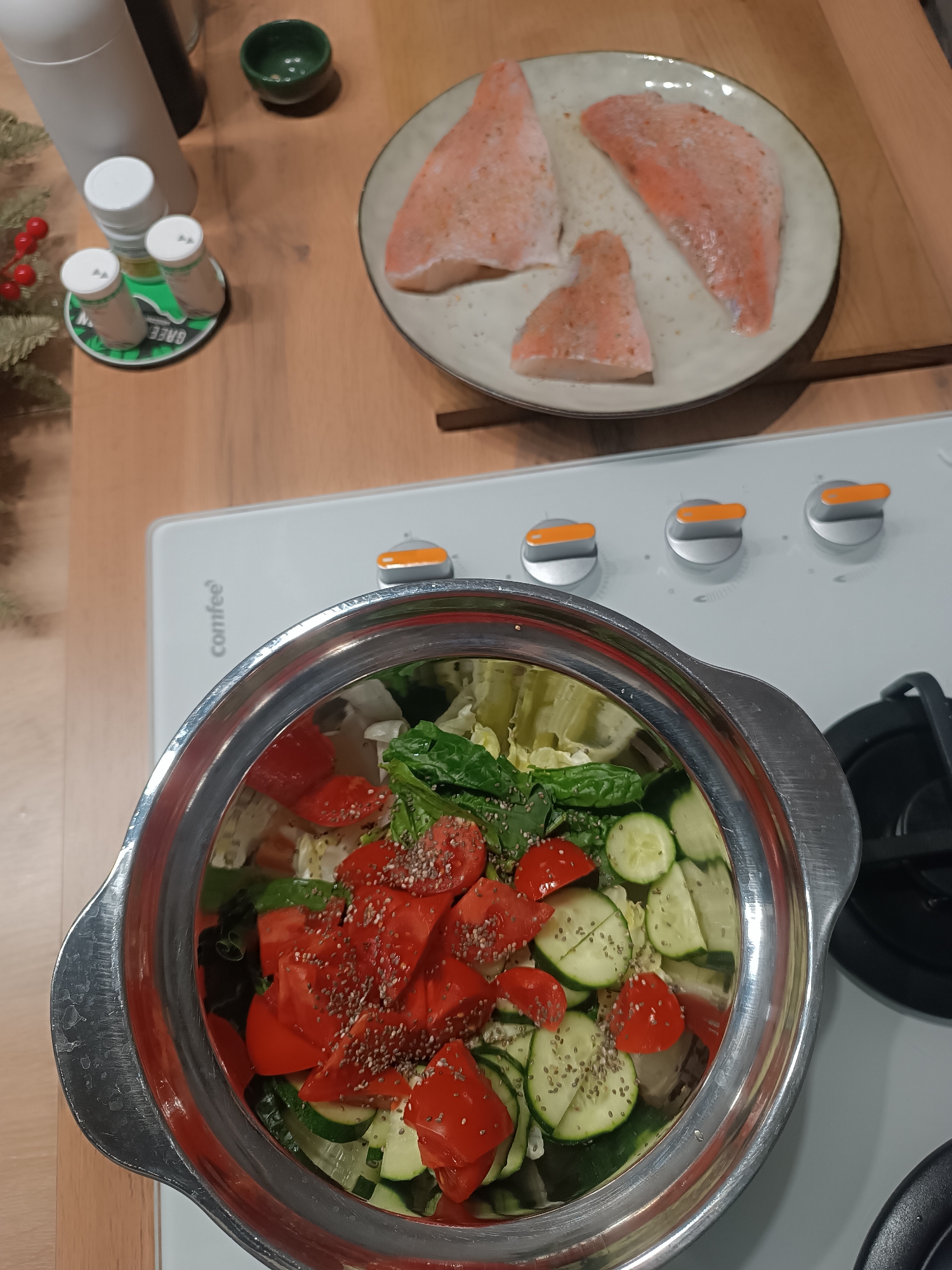 The image shows a kitchen scene with a focus on food preparation. A silver pot filled with a salad containing lettuce, tomatoes, cucumbers, and chia seeds sits on a white Comfee-branded stovetop. To the upper right, a plate holds three pieces of raw salmon, sprinkled with spices. Other elements on the wooden countertop include white and black condiment containers, a small green bowl, and what appears to be a decorative branch with red berries. The photo's setting is not Сепыч, Russia.