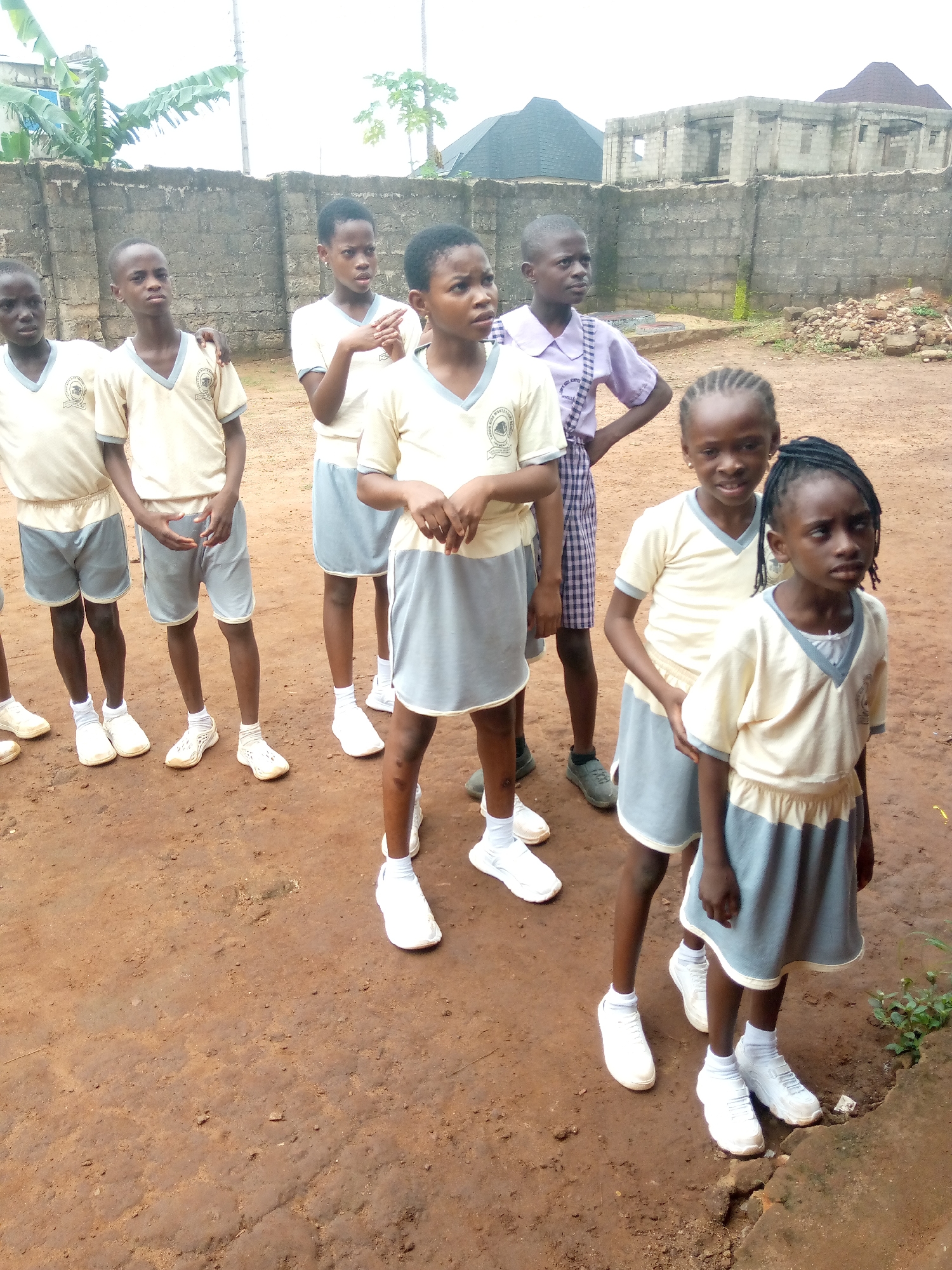The image shows a group of seven children in uniform standing on a dirt ground in front of a brick wall. The children are of varying ages, with some appearing to be younger than others. They are all wearing light-colored shirts with a V-neck collar and darker trim, paired with gray shorts or skirts. Some of the children have badges or emblems on their shirts. The environment appears to be an outdoor setting, possibly a school compound, with a rough, unpaved surface. In the background, there is a partially constructed building and some vegetation. The overall impression is one of a casual gathering or a school activity. The location in the city of Ukwu-Abwa, Nigeria, is not explicitly discernible from the visual elements alone, but it is provided as context.