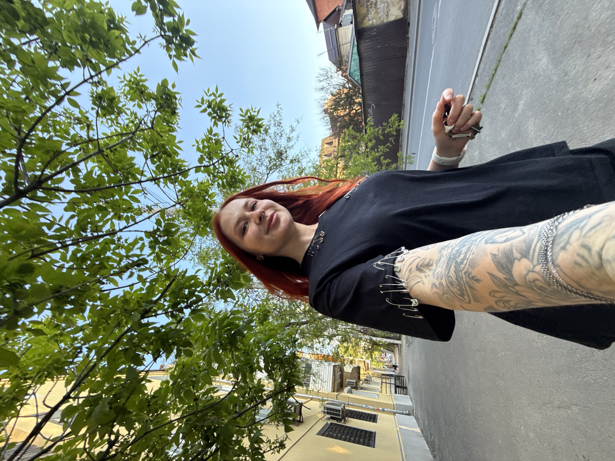 A young woman with red hair is taking a selfie outdoors in Rostov-on-Don, Russia. She is wearing a black t-shirt adorned with safety pins and has a tattoo covering her right forearm. She is holding a small white object, possibly a tissue, in her right hand. The background includes a street, buildings, and trees with green leaves. The buildings appear to be older residential structures, some with air conditioning units installed. The sky is visible in the upper portion of the frame.
