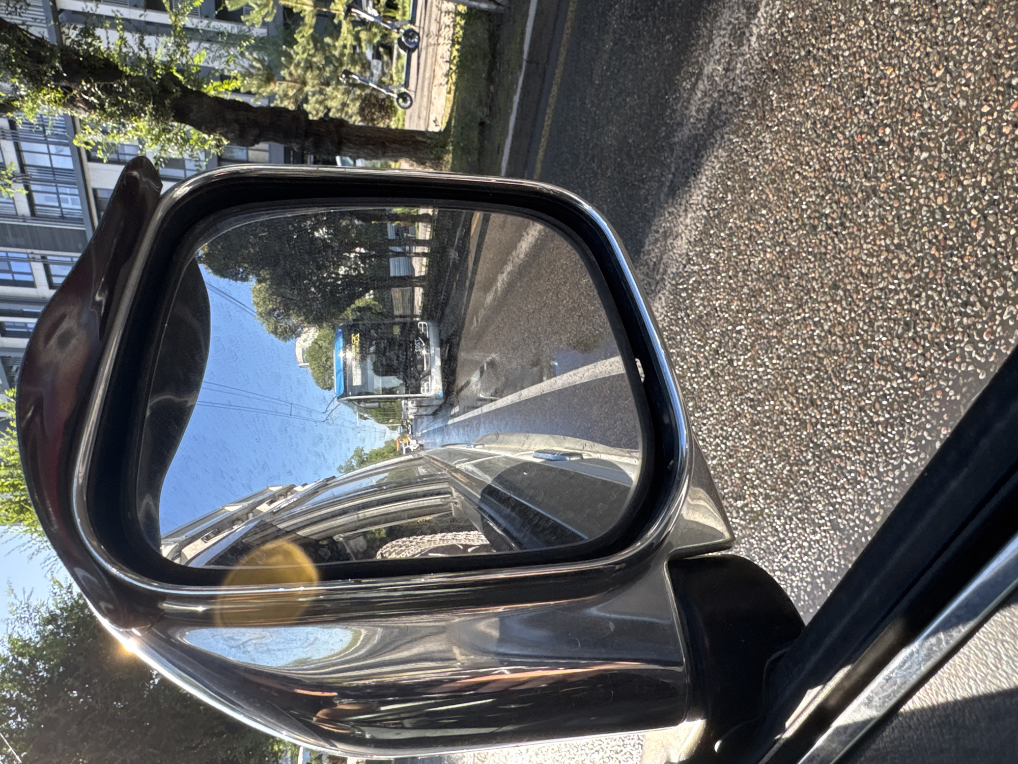 The image is a reflection in a side-view mirror of a vehicle traveling on a wet asphalt road in Almaty, Kazakhstan. The reflection shows a modern white and blue bus with the number "1303" visible on its front. The bus is on the same road, indicating it is in front of the vehicle. Trees line the roadside, and a tall, modern building with multiple windows can be seen in the background, characteristic of urban areas in Almaty. Two electric scooters are parked on the sidewalk on the right side of the reflection. The sky is clear and blue. The overall scene depicts a typical urban streetscape in Almaty during daytime.