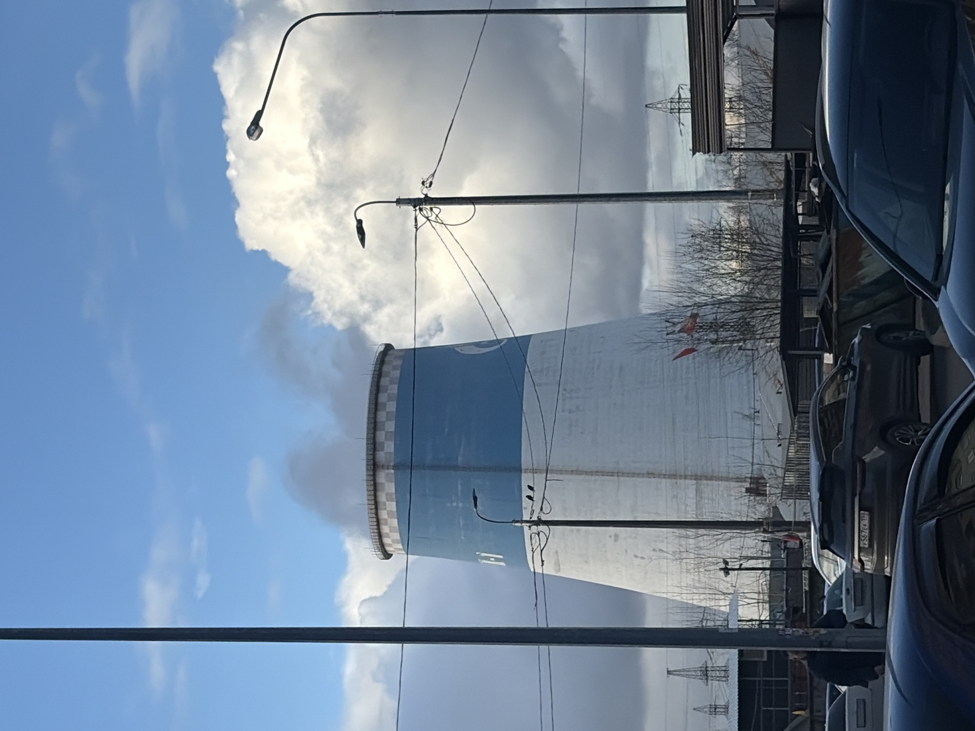 The image shows a large industrial cooling tower with a blue band at the top, emitting vapor, in the city of Saint Petersburg, Russia. The tower is situated behind a row of parked cars and streetlights. The sky is partly cloudy, with areas of blue. In the distance, transmission towers are visible. Bare trees suggest the season is late fall or winter. A utility pole is in the foreground.
