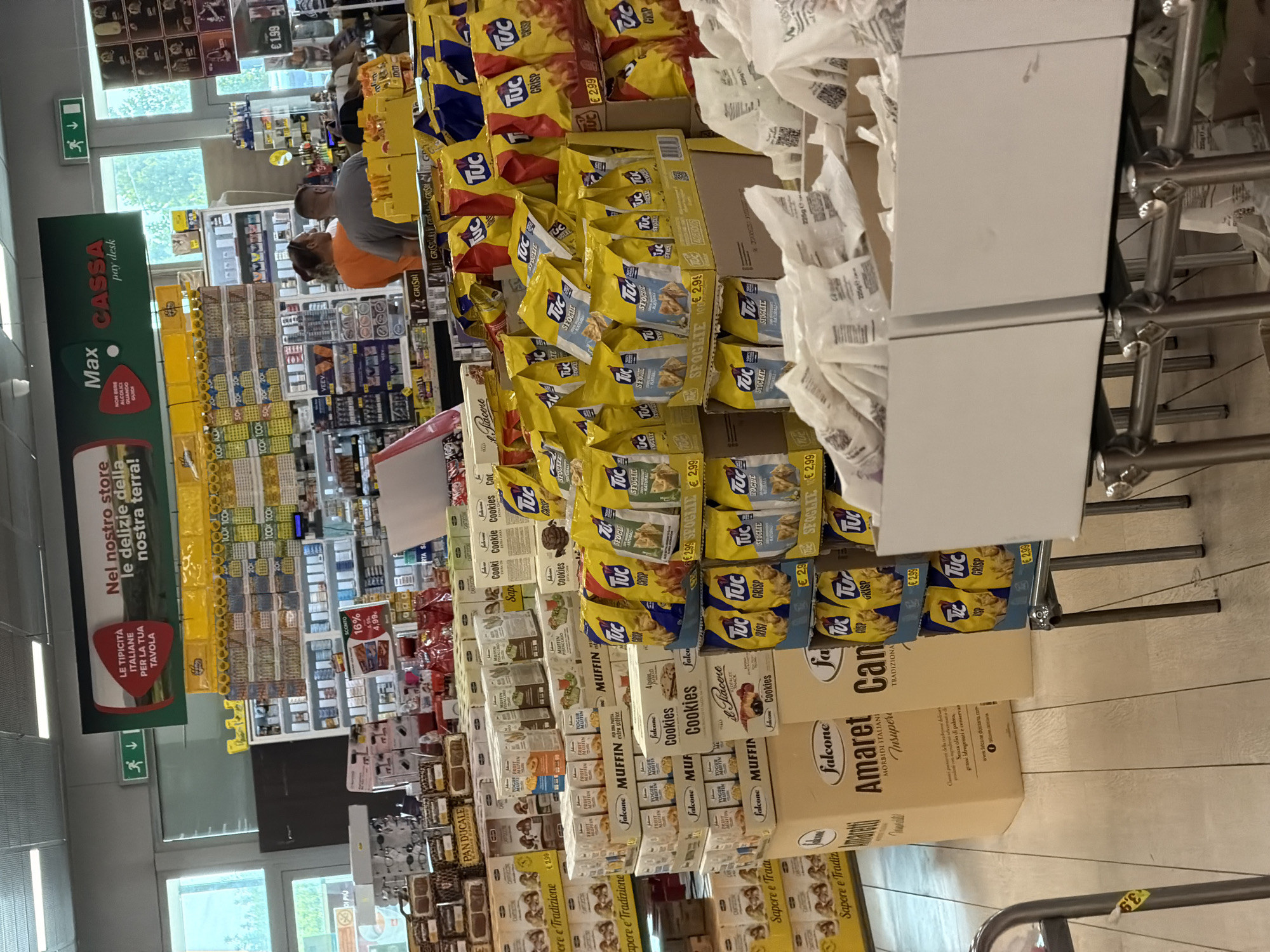 An interior shot of a supermarket displays shelves stocked with various food products, predominantly baked goods and snacks. In the foreground, large boxes of cookies and amaretti biscuits are visible, with "Amaretti Morbidi Italiani" and "Cane di Zucchero" branding on the packaging. To the right, stacks of yellow and blue bags labeled "Tuc Crisp" and "Tuc Sfoglie Naturale" are arranged on shelving units, with prices like "€ 2.99" displayed. In the background, shelves are filled with packaged goods, including what appears to be tobacco products, with signage indicating "Max CASSA pay desk". Two individuals, a man in an orange shirt and another in a grey t-shirt, are seen standing near the checkout area. The overall environment suggests a retail setting within a supermarket in Due Carrare, Italy, given the presence of Italian product labeling and the "CASSA" (cashier) signage common in Italian stores.