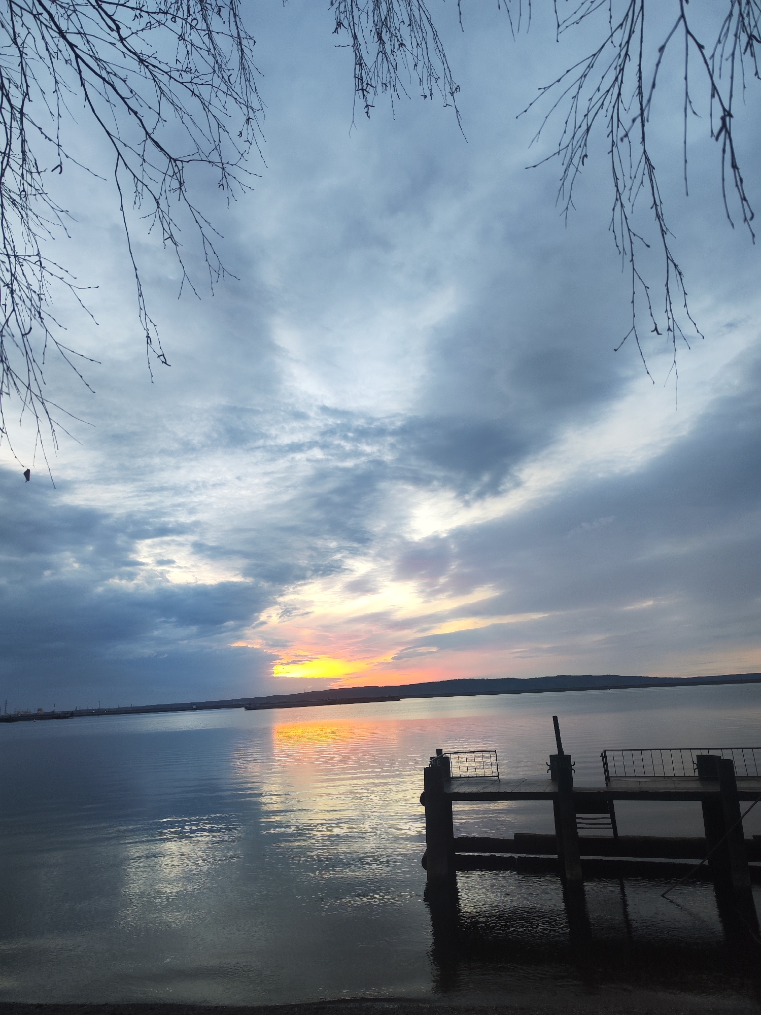 The media file presents a serene twilight landscape over a wide body of water, likely a large lake or river, situated in Chaikovsky, Russia. The scene captures either a sunrise or sunset, characterized by a vivid strip of orange, yellow, and subtle pink light radiating from the horizon. This bright band contrasts sharply with the expansive, overcast sky, which is filled with layers of grey and bluish-grey clouds.
Bare tree branches, possibly birch, with visible buds or catkins, gracefully frame the top sections of the image, silhouetted against the cloudy sky. Their intricate patterns add depth to the foreground. The water's surface is calm, mirroring the dramatic sky with both the fiery hues of the horizon and the cooler tones of the overhead clouds creating a rich reflection.
In the lower right portion of the frame, a dark, weathered pier or dock extends into the water. This structure features simple railings and supports, casting a dark reflection on the water below. Across the expanse of water, a low-lying distant shoreline is visible. On the far left of this distant landmass, faint outlines of what appear to be industrial structures or buildings can be discerned.
The overall mood is tranquil and contemplative, emphasizing the natural beauty of the moment. There are no people visible, and no specific activity is taking place. No discernible text appears in the image.