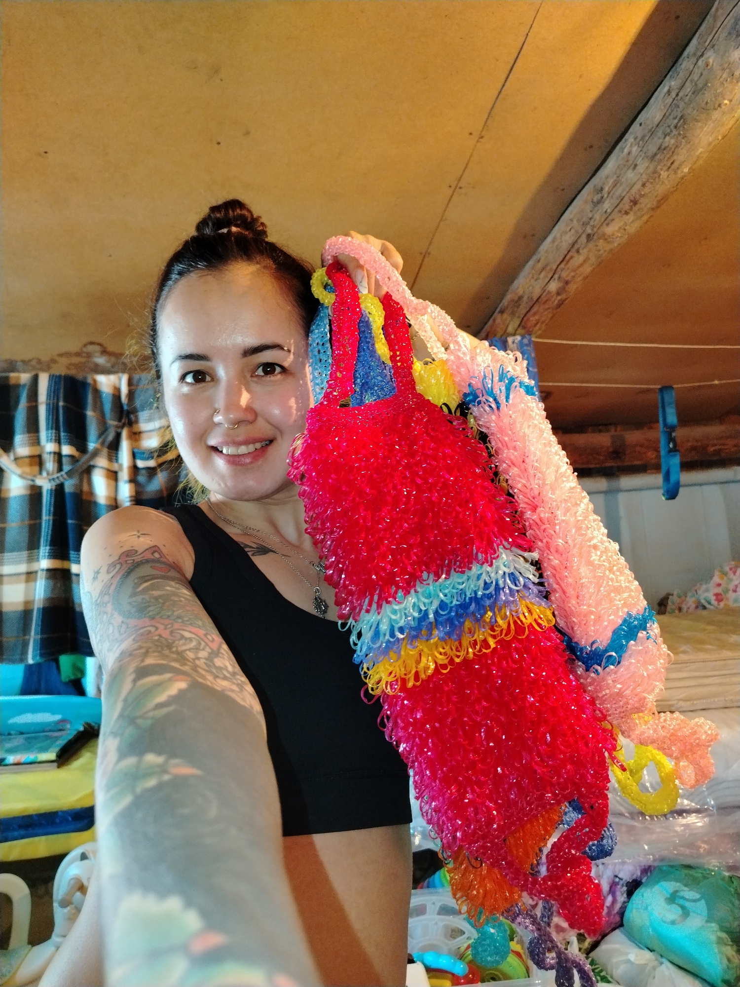 A female individual is depicted indoors in Гожан, Russia, posing for a selfie. She is smiling directly at the camera, with her head slightly tilted. Her dark hair is pulled back into a bun. She wears a small gold nose ring (septum piercing) and a silver necklace with a pendant. Her visible left arm is heavily tattooed with colorful patterns. She is wearing a black sleeveless top. With both hands, she holds up several brightly colored, textured garlands, possibly made from woven plastic or elastic bands. The most prominent garland is red with horizontal stripes of blue, yellow, and orange; another is light pink with blue accents.
The background features a light brown, possibly wooden, wall or ceiling. A rustic wooden beam is visible diagonally in the upper right. To the left, a blue and beige plaid fabric hangs, possibly a curtain. A thin white line, resembling a clothesline, spans the mid-ground, from which a blue strap-like object hangs. The lower right background contains various items, including more colorful plastic objects and clear plastic bags, suggesting a storage or craft area.