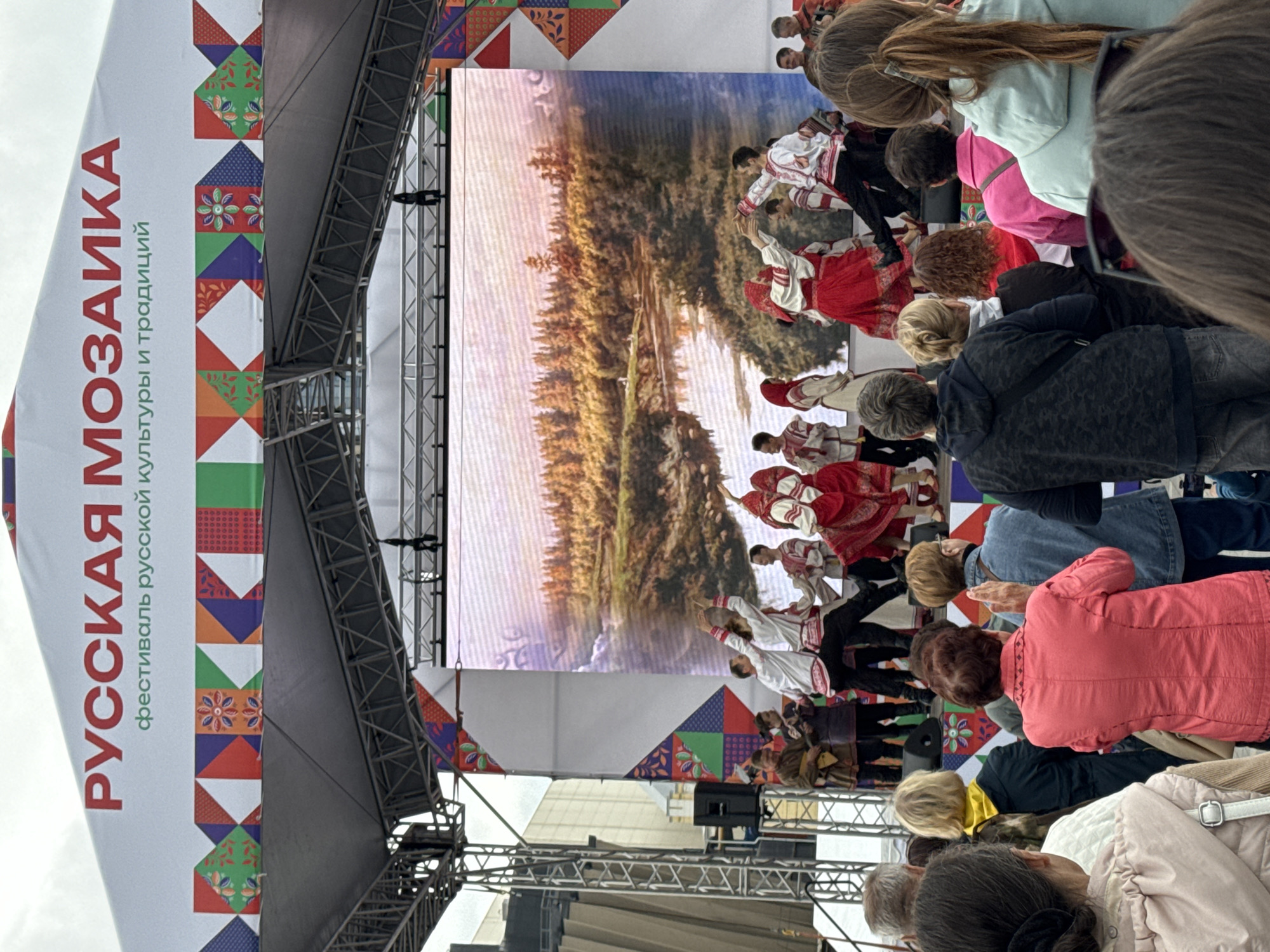 This image depicts an outdoor cultural festival, identified by the banner as the "Russian Mosaic" festival of Russian culture and traditions, held in Perm, Russia. A large, temporary stage structure occupies the center of the frame, topped with a white banner displaying red Cyrillic text "РУССКАЯ МОЗАИКА" (Russian Mosaic) and smaller green text "фестиваль русской культуры и традиций" (festival of Russian culture and traditions). Beneath the text, a colorful border composed of geometric and floral patterns runs horizontally along the banner.
On the stage, a prominent LED screen serves as a backdrop, displaying a realistic landscape image of a river flowing through a dense forest under a slightly overcast or hazy sky. In front of the screen, a group of approximately ten performers is engaged in a folk dance and musical performance. Male dancers wear white shirts with red embroidery and dark trousers, while female dancers are dressed in red skirts, white blouses, and red traditional headwear. They are captured in dynamic poses, characteristic of Russian folk dance, with arms raised and legs in motion. To the left of the dancers, two musicians, also in traditional attire, are visible; one plays a balalaika, and another holds a stringed instrument. A black speaker unit is positioned on the stage floor to the left.
The foreground is filled with the backs of a diverse audience, numbering at least twenty individuals, observing the performance. Spectators are dressed in casual attire, including jackets, hoodies, and various tops. One woman in the center-left foreground wears a prominent red jacket and appears to be clapping. The scene is illuminated by natural, diffused daylight under an overcast sky. Partially visible behind the stage structure on the left is a modern building with a textured, possibly metallic, facade.