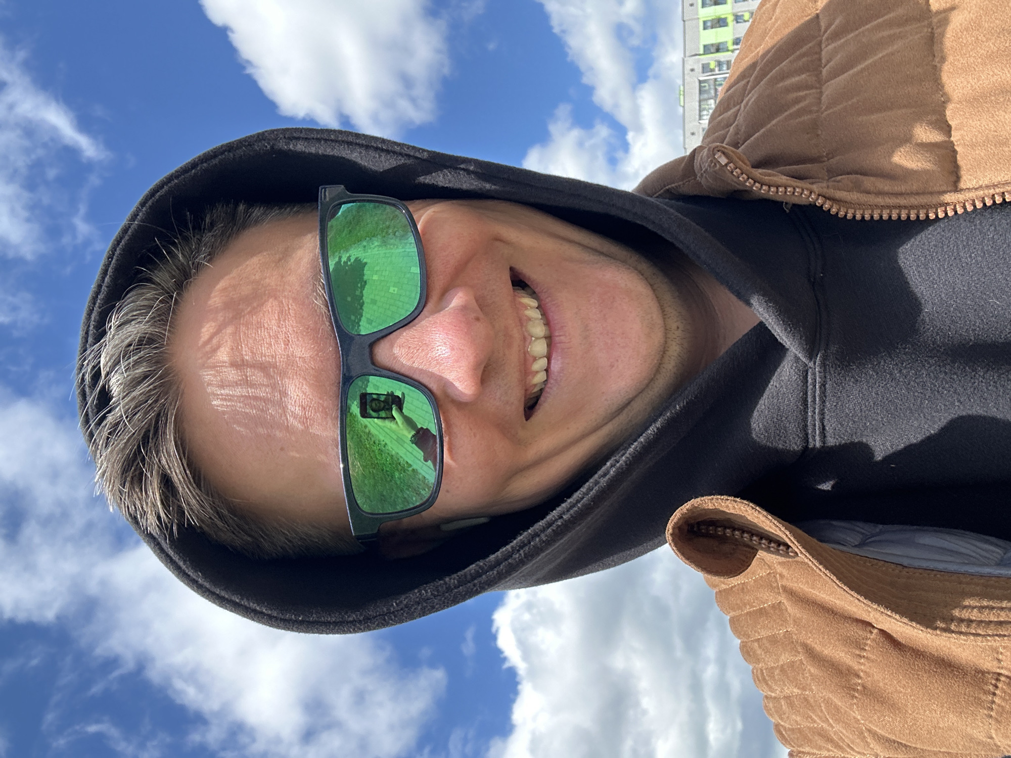 A low-angle selfie shows a man wearing green-tinted sunglasses, a black hooded sweatshirt, and a brown quilted jacket. He is smiling, and his head is tilted. The reflection in his sunglasses shows a person holding a phone. The background consists of a blue sky with scattered white clouds and a partially visible building with light green siding. Based on the query, the location is Бараўлянскі сельскі Савет, Belarus.