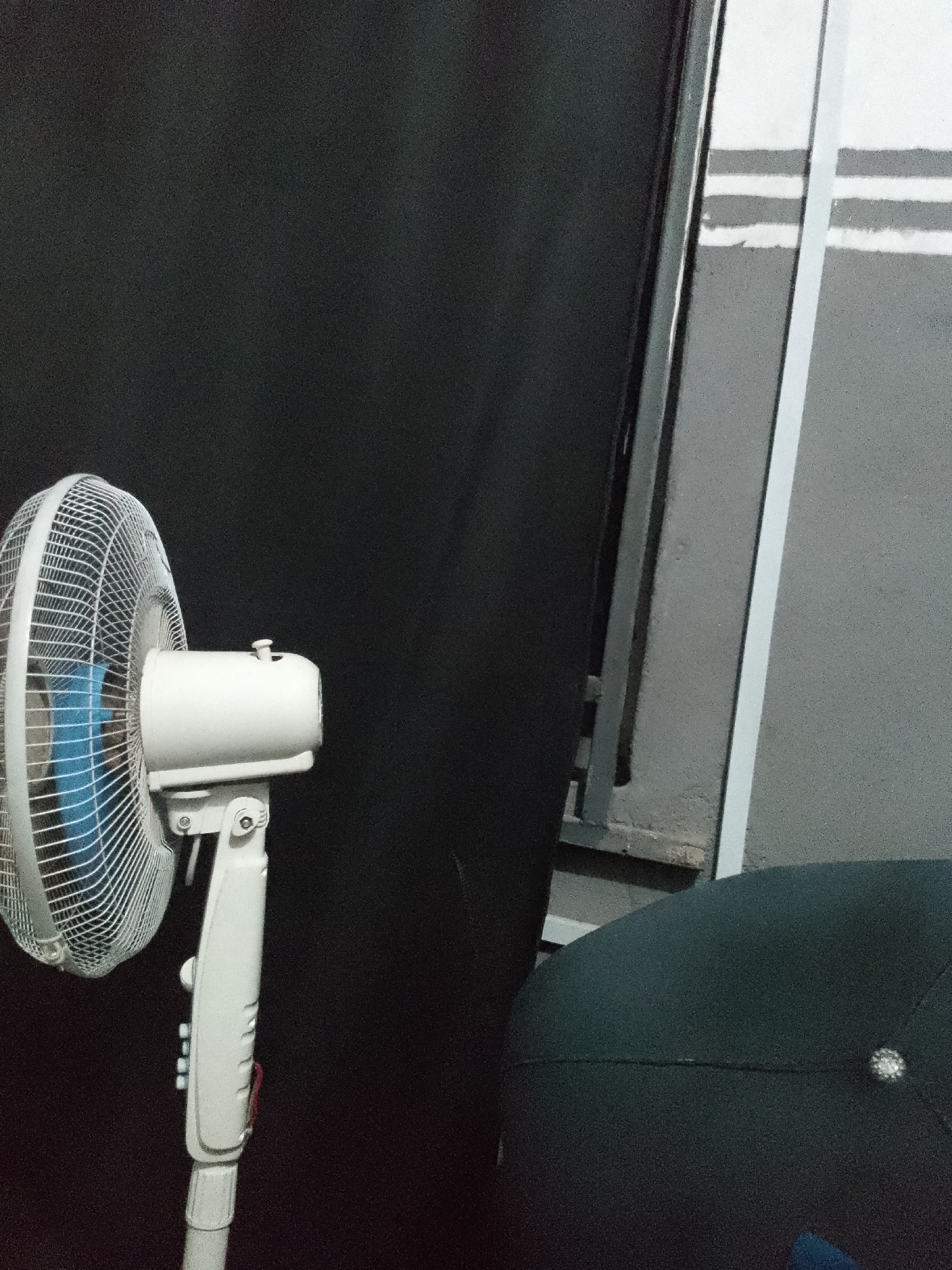 An indoor, low-light scene shows a white standing fan on the left, partially obscuring a dark, textured background that appears to be fabric or a wall. The fan has a silver grill and a blue fan blade visible within. To the right of the fan is a portion of a grey, textured wall with horizontal white stripes. In the foreground, the top of a dark, round object, possibly a cap or a hat, is visible. A thin, vertical white strip runs from the top edge of the frame down the right side, adjacent to the grey wall. The scene suggests an interior setting, possibly a room or studio, with no visible indication of a specific city or geographical location.
