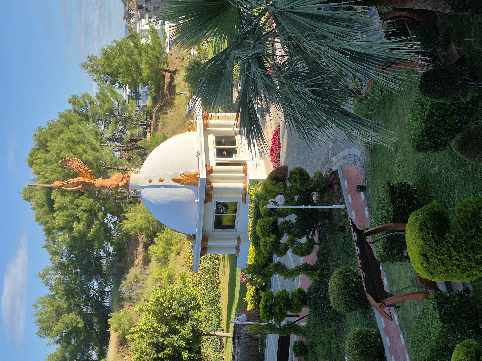 The image shows a white domed structure with a gold winged figure on top, located in a garden setting with trees, bushes, and a brick path. The structure has windows and decorative gold accents. In the background, there are trees and buildings, and the sky is visible. The location is Alanya, Turkey. A palm tree is on the right side of the image and a bench is in the foreground.