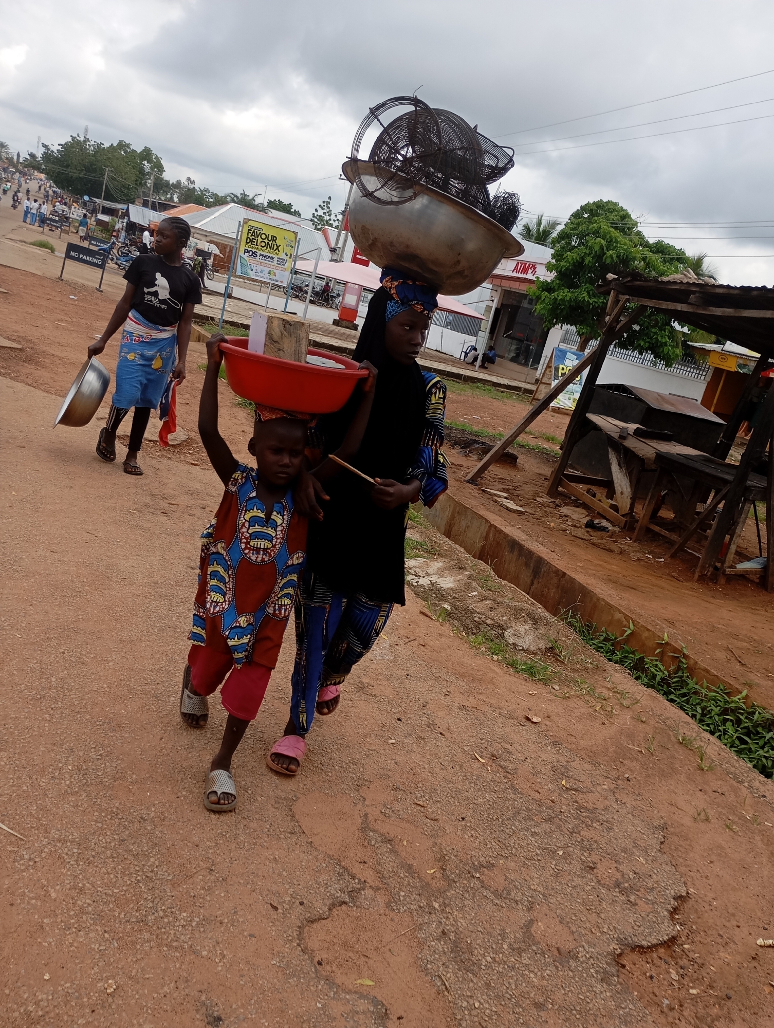 Two females, one adult and one child, are walking on a dirt road in Shendam, Nigeria. The adult female, on the left, is wearing a black t-shirt and a blue and white patterned skirt. She is holding a silver bowl in her left hand. The child, on the right, is wearing a colorful patterned tunic and red pants, and is carrying a red bowl on her head. The adult female is looking to her left, while the child is looking forward. In the background, there are buildings, trees, and other people walking and riding motorcycles. The sky is cloudy. The scene appears to be a street market or a busy road in a town.