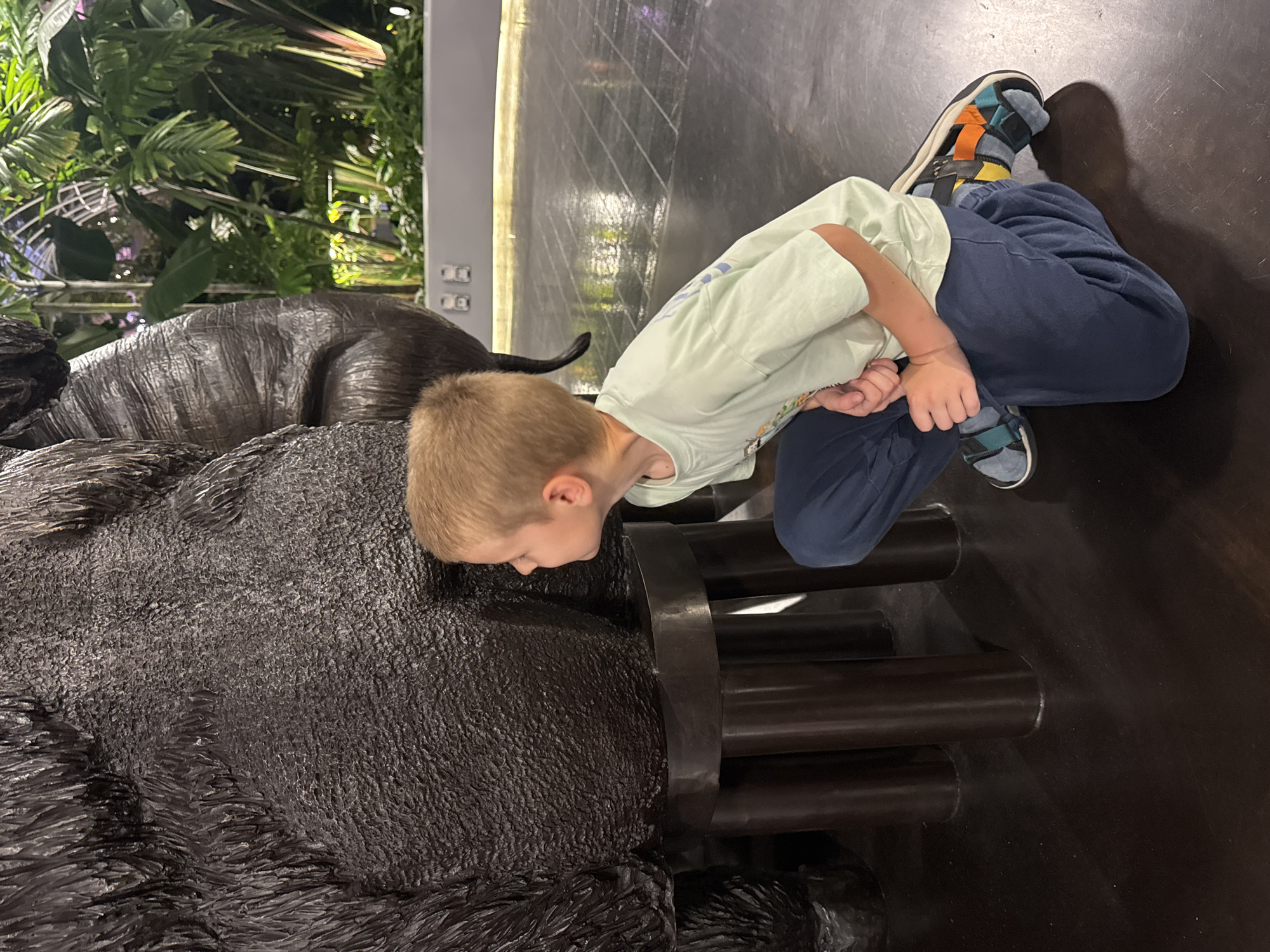 A young boy, with blond hair and wearing a light green t-shirt and navy blue pants, crouches down to examine the textured backside of a large, dark, gorilla statue in the Jewel Changi Airport, Singapore. The statue is supported by cylindrical metallic legs. In the background, lush green tropical plants and artificial lighting are visible within a modern, enclosed environment. The floor is a dark, reflective surface with a grid pattern.