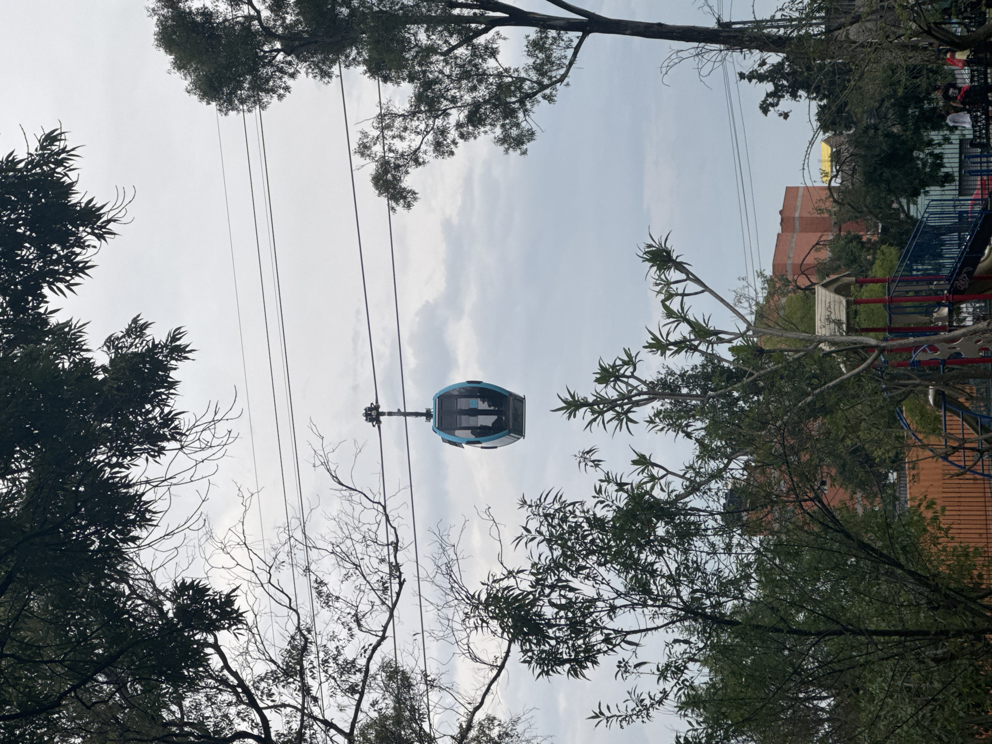 The image shows a cable car suspended in the air. It is framed by trees, with a cloudy sky in the background. A building and a playground structure are partially visible behind the trees on the right side of the image. The location is the city of Unknown, Unknown.