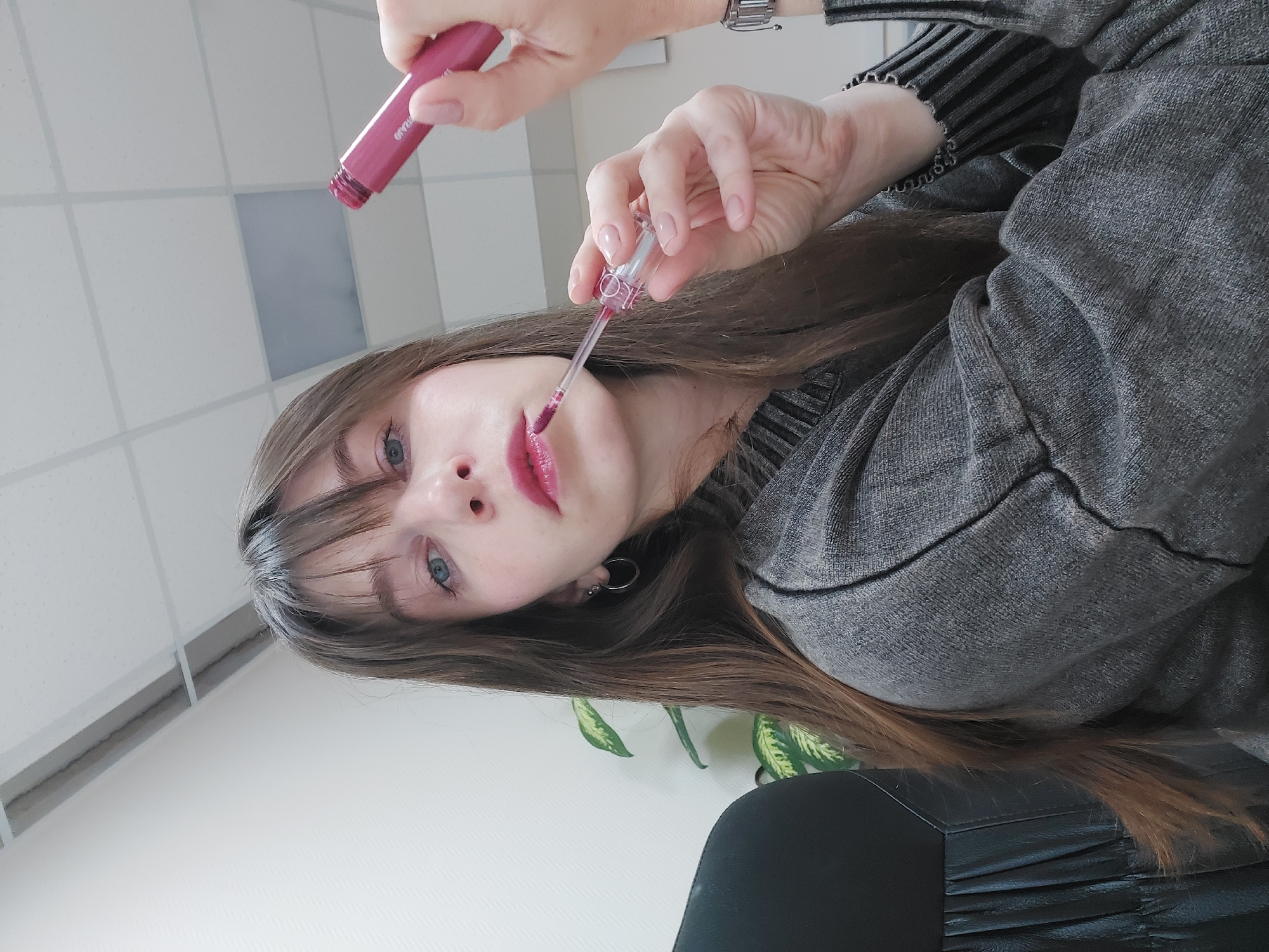 A woman with long brown hair and bangs is applying lip gloss. She is wearing a grey sweater. The background includes a white ceiling with recessed lighting, a white wall, a plant with green and yellow leaves, and a black object that may be a chair. While the location is claimed to be Chaikovsky, Russia, there is no verifiable evidence in the image to confirm that location.