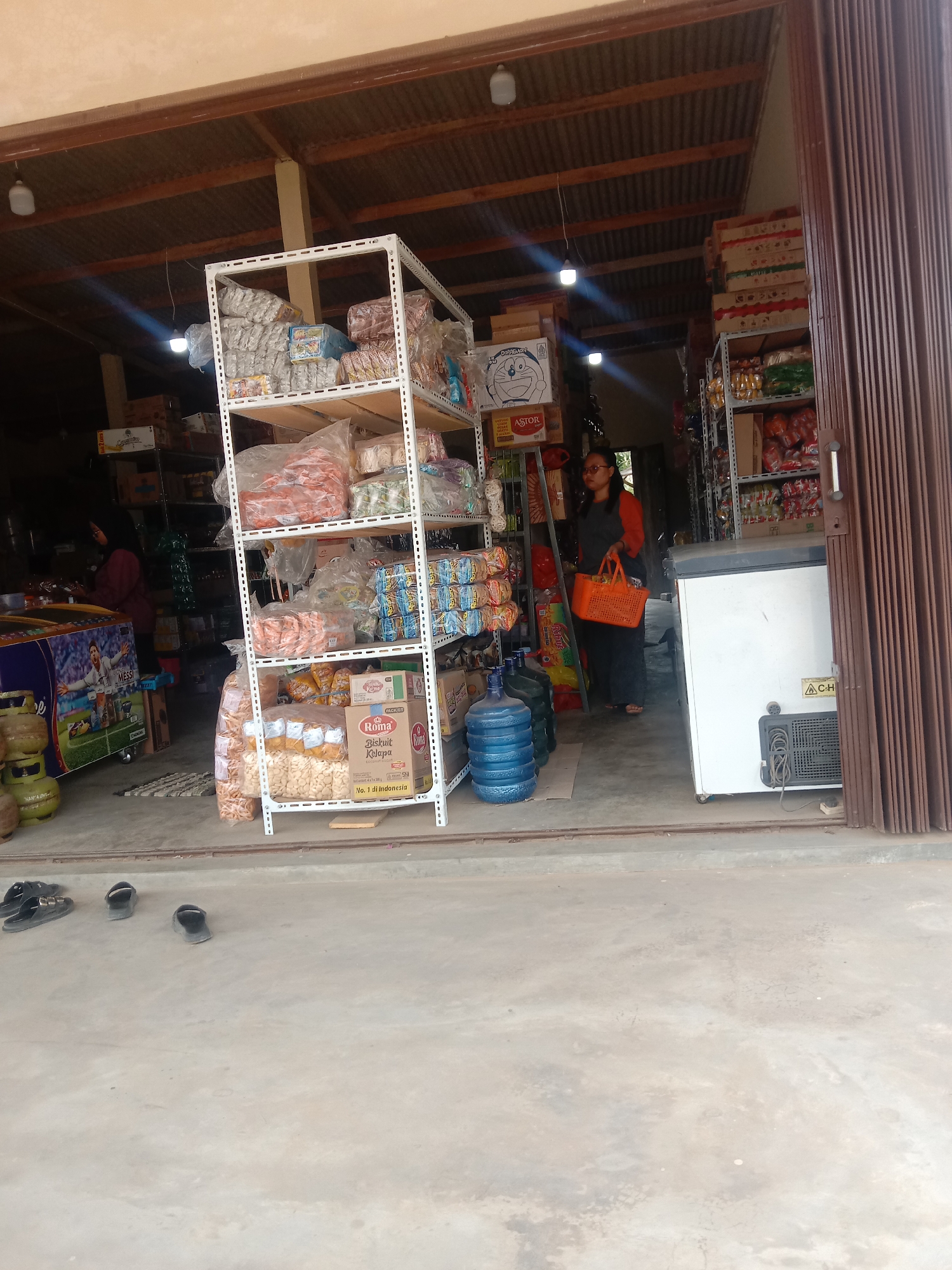 The image shows the entrance of a small convenience store, possibly located in Utama Jaya Mataram, Indonesia. The store is stocked with various goods, displayed on white metal shelves. In the foreground, on the concrete floor, there are several pairs of black sandals. Inside the store, a woman wearing glasses, a red long-sleeved shirt, and a dark skirt is standing and holding an orange basket. She appears to be a customer or an employee. A white chest freezer with a dark gray vent is visible on the right side of the entrance. Several large blue water jugs are stacked near the freezer. The store's ceiling is corrugated metal, supported by wooden beams. The overall impression is that of a typical small neighborhood store.