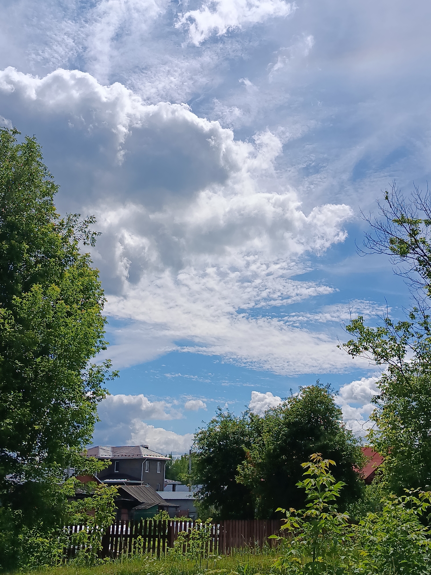 The image depicts a rural landscape in the city of Verreya, Russia, under a bright, partly cloudy sky. In the foreground, lush green foliage and trees frame the scene, with a wooden fence running horizontally across the lower portion of the image. Behind the fence, several houses are visible, with varying rooflines and architectural styles, suggesting a residential area. The sky is a prominent feature, with large, billowing white clouds interspersed with patches of clear blue. The overall impression is one of a serene, summer day in a quiet, rural setting.