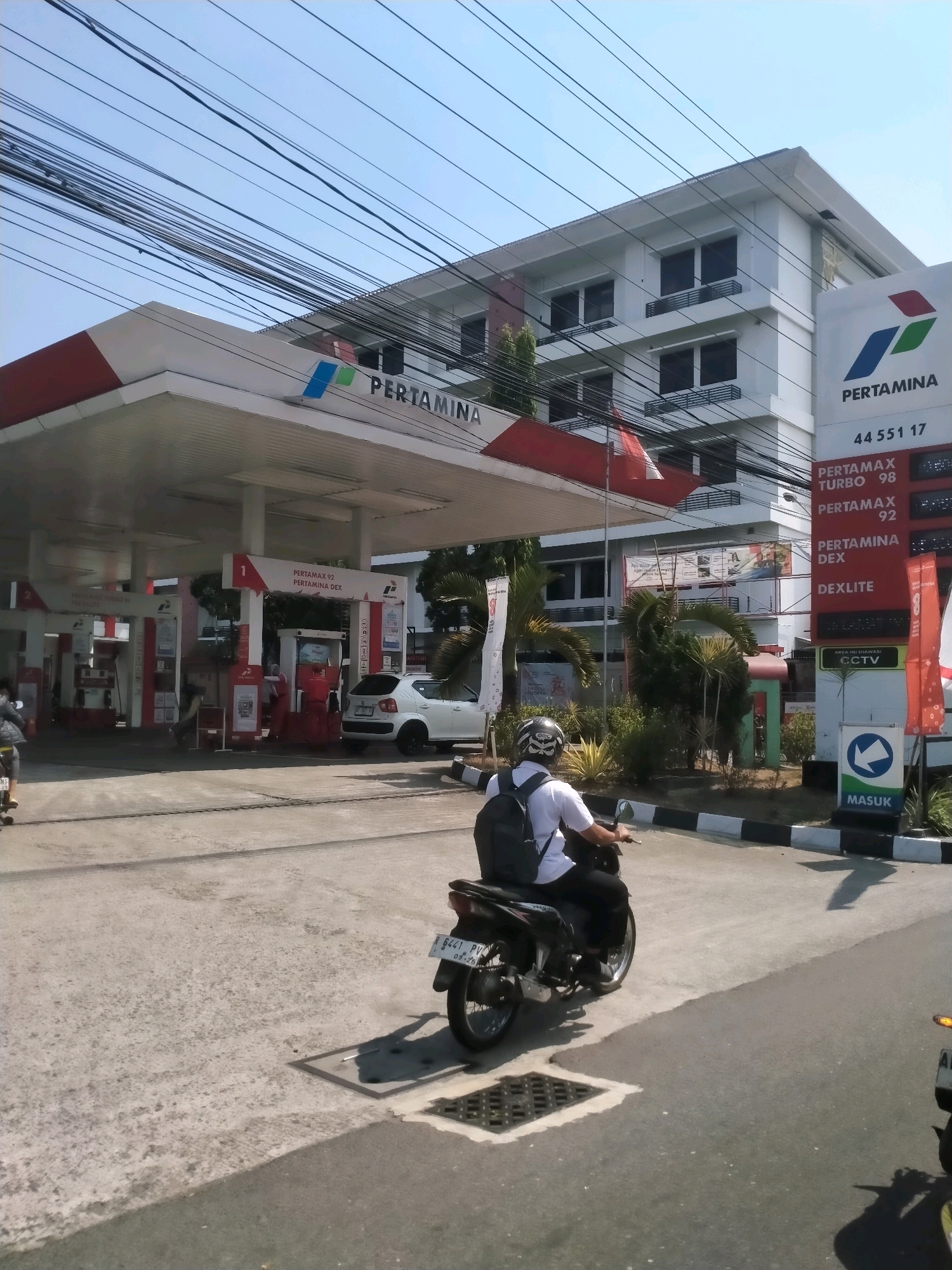 A Pertamina gas station is depicted in Yogyakarta, Indonesia. The scene is observed from a street-level perspective, with a clear blue sky above and overhead power lines crisscrossing the frame. The gas station features a white canopy with red and white trim, under which several fuel pumps are visible. A multi-story building with white walls and dark windows stands behind the gas station. A man wearing a white t-shirt, black pants, and a backpack is riding a motorcycle towards the gas station, his face obscured by a helmet. Other vehicles, including a white car, are present at the station, and a sign with the text "MASUK" (meaning "enter" in Indonesian) and a directional arrow is visible. The ground is paved with concrete.