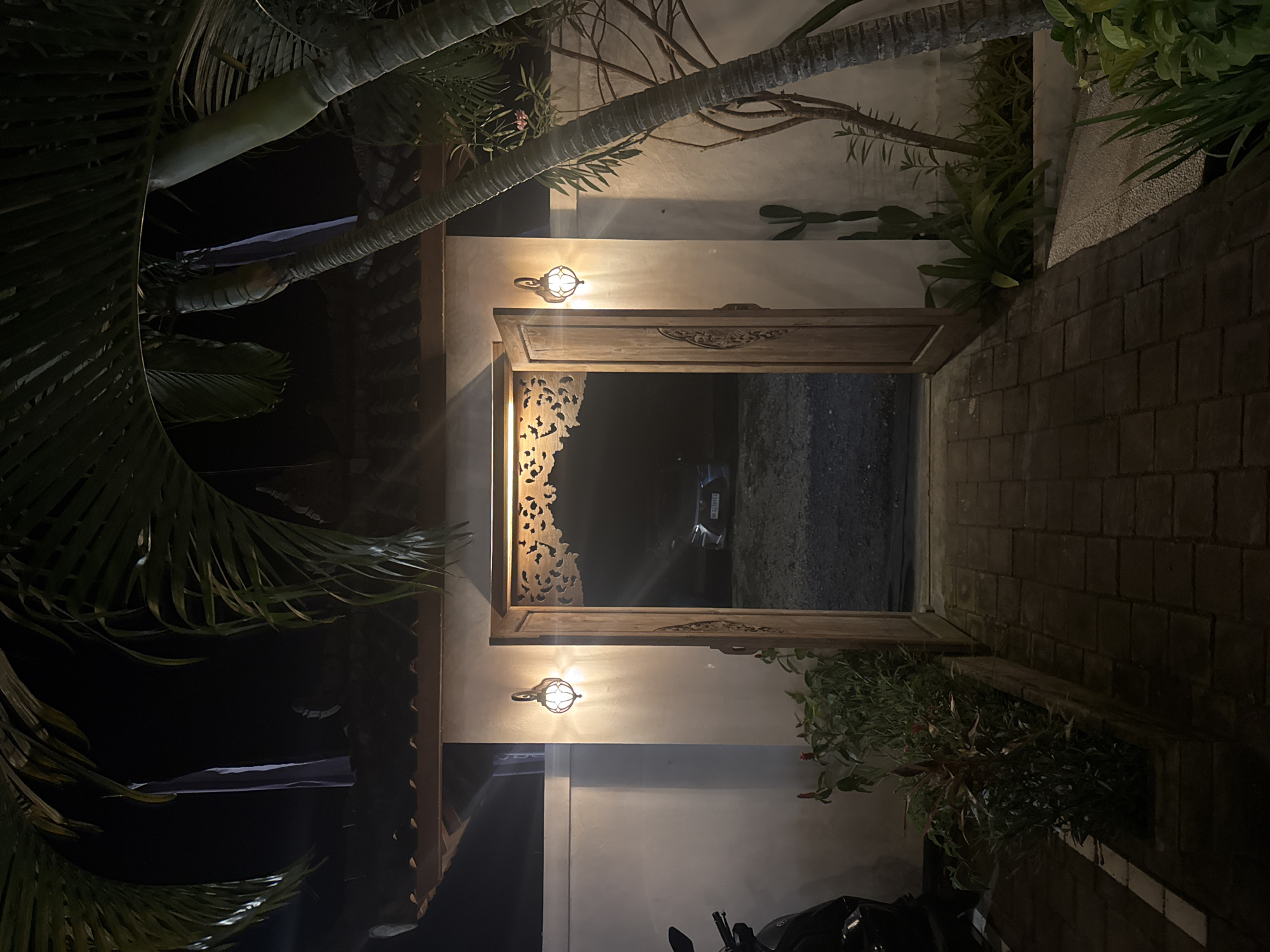 The image depicts an outdoor scene in Kuta Selatan, Indonesia, at night. A doorway frames a view into a dark area where a car is parked. The doorway itself is wooden and ornately carved. Two sconce lights are mounted on the wall flanking the doorway, providing illumination. Tropical plants and palm trees surround the structure. The walkway leading to the doorway is paved with bricks. A potted plant sits on the walkway edge near the bottom right corner. A motorcycle is also partially visible in the bottom right corner.