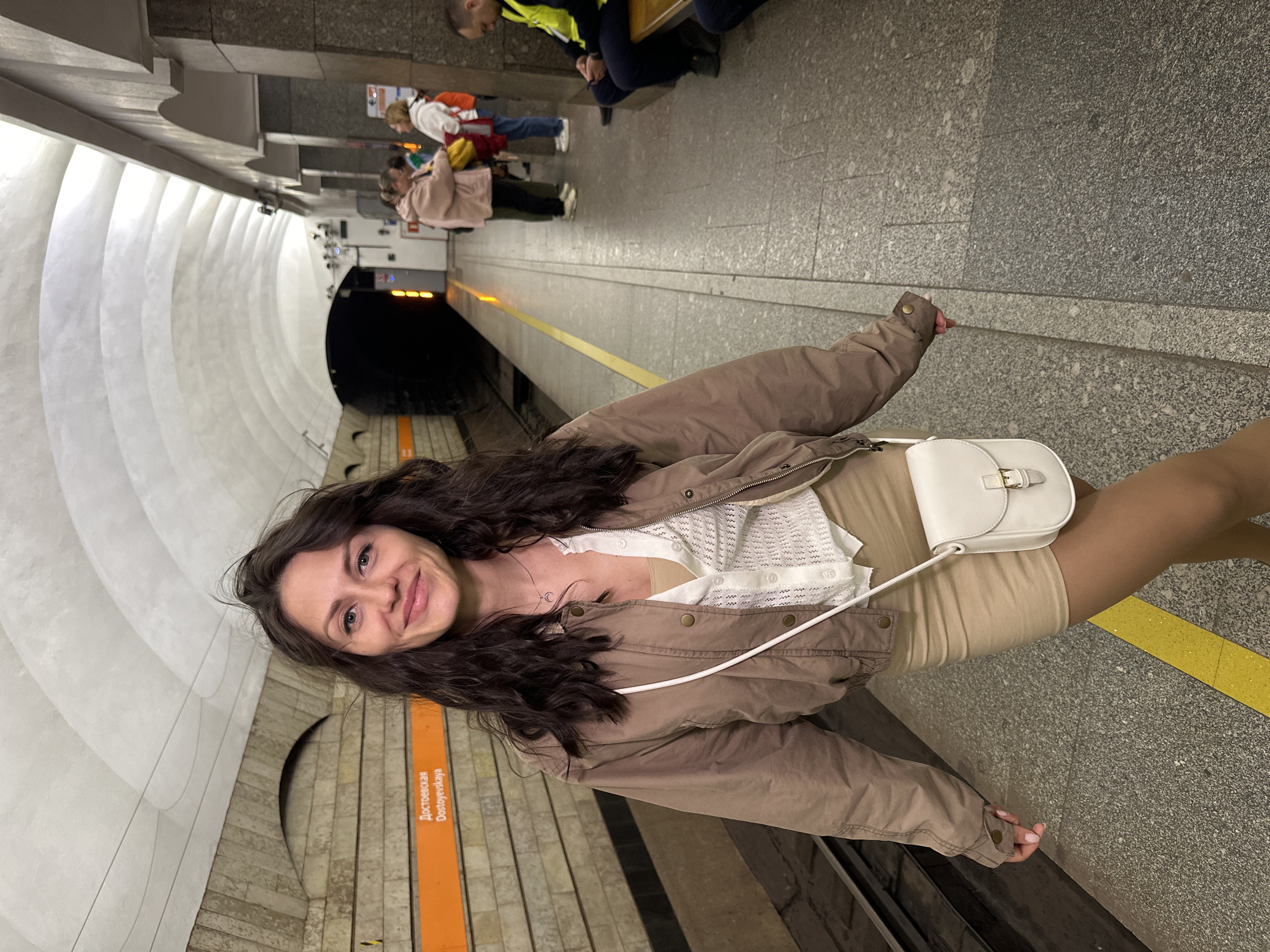 A woman is standing on a subway platform in Saint Petersburg, Russia, likely at Dostoyevskaya station, indicated by the orange sign. She is wearing a beige skirt, a white ruffled cardigan over a beige top, and a brown bomber jacket. A small white purse is slung across her body. Her dark hair is down, and she is smiling at the camera. In the background, other commuters are visible, some waiting on the platform, and a train tunnel is visible in the distance. The platform features typical subway station architecture with arched ceilings and granite-clad walls.
