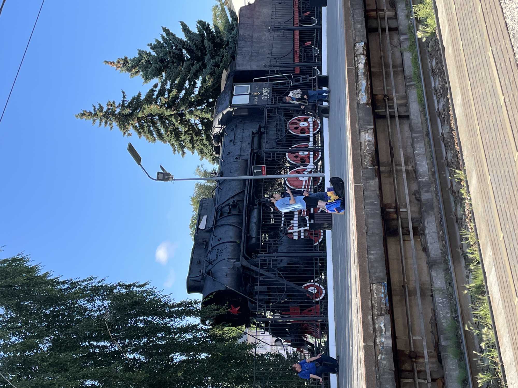 The image shows a black steam locomotive, preserved as a monument, on display at a train station platform in Vladimir, Russia. The locomotive is large and imposing, with red accents on its wheels and a red star emblem on its front. Several people are present on the platform, some standing and observing the train, while others are seated. One person in a blue shirt is sitting on a bench to the left. Two men in casual attire are standing near the front of the locomotive, and another man in a patterned jacket is near the side of the train. A child in a blue shirt is sitting on the platform next to a backpack. The platform itself is made of concrete and runs alongside the train tracks. The background features trees, a lamppost, and a clear blue sky. The scene appears to be a sunny day, with good visibility.