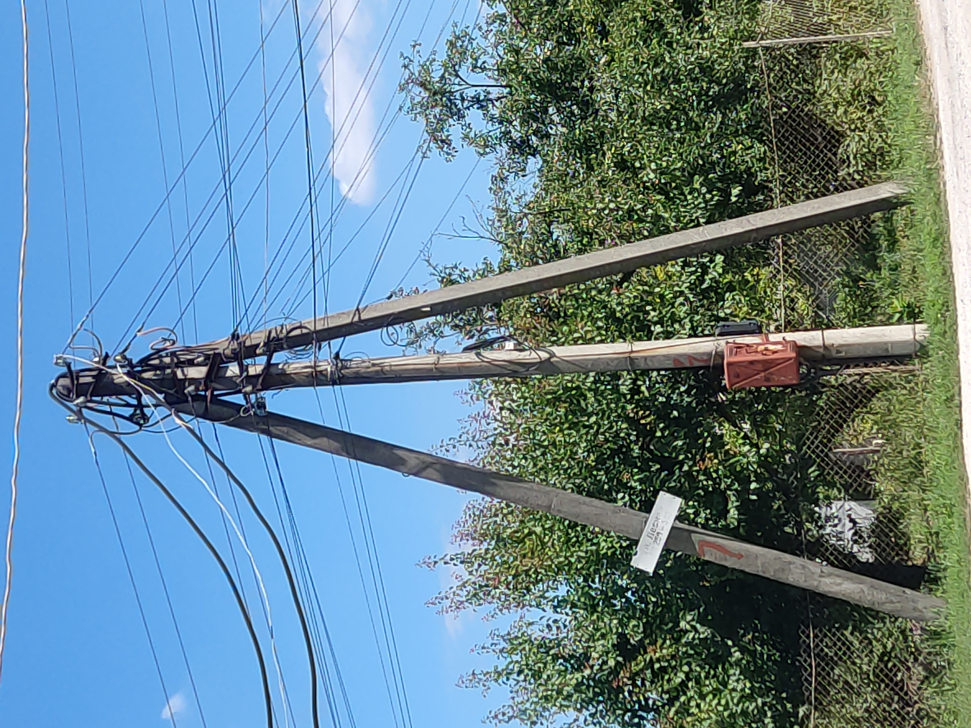 This is a medium-distance, eye-level, outdoor, daylight, static shot of a triangular power pole against a bright blue sky with some scattered clouds. The power pole is constructed of three concrete supports meeting at the top, where electrical wires are strung and connected to a central junction box with numerous insulators and wiring. A red electrical box is mounted on one of the concrete supports about a third of the way up from the base. Attached to another concrete support, lower down and at an angle, is a white rectangular sign with black lettering. A red number "7" is painted on the same support below the sign. Behind the power pole, a dense line of green trees with some reddish fruit is visible, with a chain-link fence in front of the trees. The ground in the foreground is a dirt or gravel road. The scene appears to be in Radisne, Ukraine, based on the presence of the sign which reads "с. ЛЕСИ" and a number that might be a street or property designation.