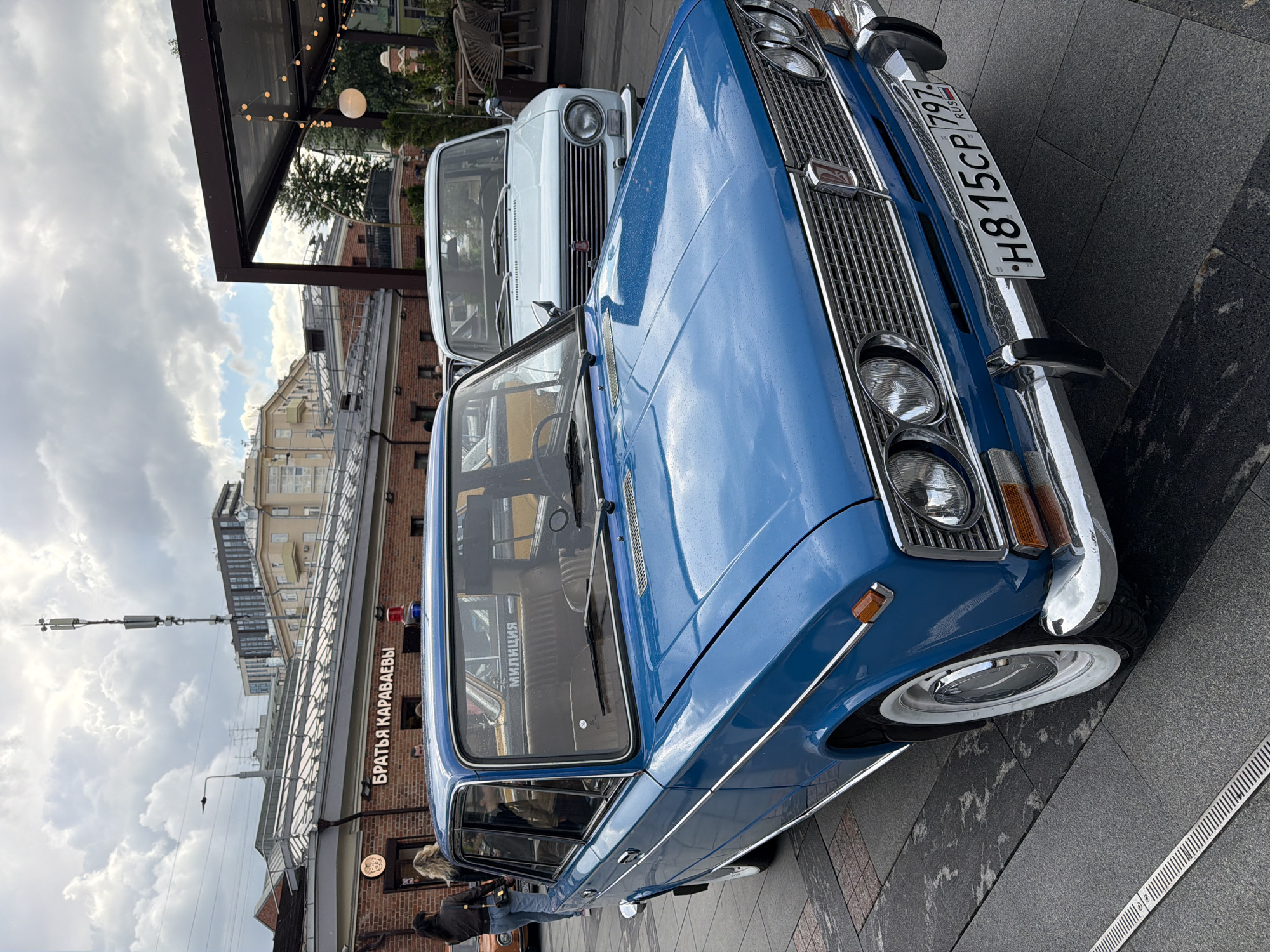 The image shows two vintage cars parked on a city street. In the foreground is a blue Lada 2106 sedan, its license plate visible and reading "H815CP 797 RUS". Behind it is a white Volga GAZ-24. The cars are parked outside a building with a sign reading "БРАТЬЯ КАРАВАЕВЫ" (Brothers Karavaev), suggesting the location is near a restaurant or business with that name. The surrounding environment includes urban architecture, street paving, and overcast skies. A person is partially visible near the blue car. A weather station is affixed to the building on top.