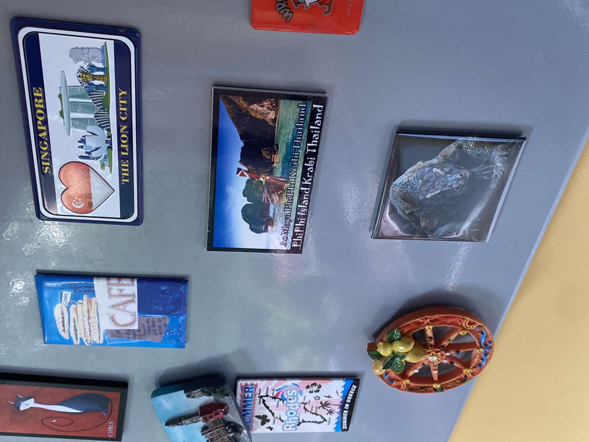 The image shows a collection of refrigerator magnets on a grey magnetic surface. The magnets depict various tourist attractions and cultural symbols. From left to right, the top row features a magnet with a black cat silhouette and the text "CYPRUS". Next to it is a blue magnet with a stack of food items and the word "CAFE". To the right of that is a rectangular magnet with an illustration of Singapore's skyline and the text "SINGAPORE THE LION CITY".
The middle row contains a magnet with a picture of boats in a tropical cove, with text that reads "Ao Maya Phi Phi Krabi Thailand Phi Phi Island Krabi Thailand". To the right of this is a red magnet with an anthropomorphic character and some text, but it is partially obscured.
The bottom row includes a magnet with a colorful, decorative wheel adorned with fruit and intricate patterns. Below this, a magnet displays a close-up image of a Komodo dragon. There is also a magnet with a map of Rhodes and palm trees, with the text "Rhodes" and "SUMMER IN GREECE". Another magnet, partially visible on the far left, shows a cityscape.
The background is a plain grey surface, likely a refrigerator door. The lighting is even, suggesting an indoor environment. There are no people visible in the image. The image does not contain any information about the city of Wrocław, Poland.
