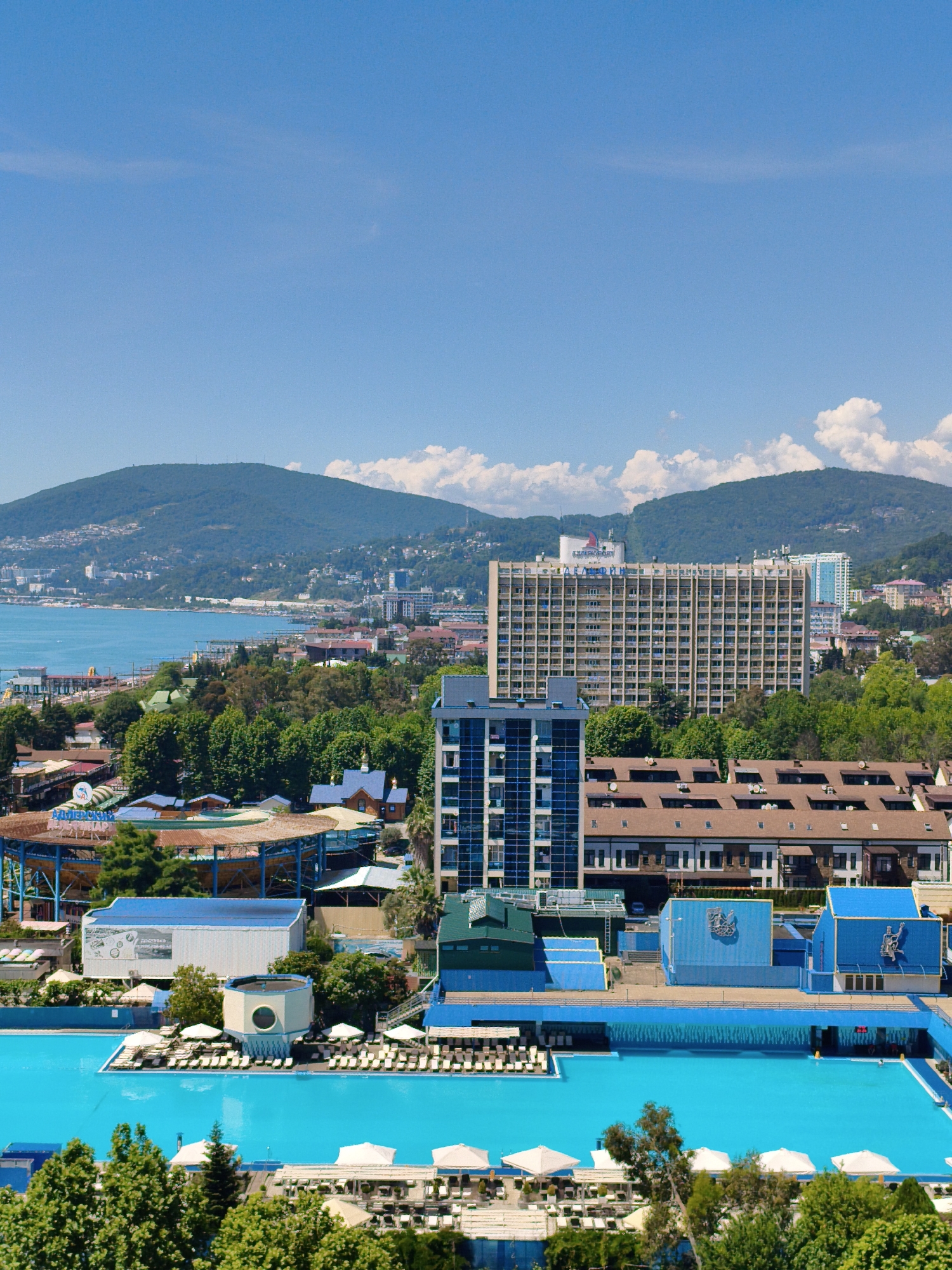 This image captures a panoramic view of a resort area in Sochi, Russia, on a clear, sunny day. The scene is dominated by a large, bright blue swimming pool in the foreground, surrounded by rows of white lounge chairs and umbrellas, suggesting a recreational atmosphere.
Behind the pool, several buildings are visible. To the left, a blue and white structure with a large banner displays text in Cyrillic. Further back, a large, multi-story hotel with numerous balconies stands prominently, with more buildings and greenery cascading up the hillsides. In the distance, lush green mountains rise against a clear blue sky dotted with a few fluffy white clouds. The sea is visible in the background, calm and blue.
The overall impression is one of a vibrant, summer resort town. The bright sunlight and clear skies indicate fair weather, ideal for outdoor activities. While no people are clearly discernible in the foreground, the presence of lounge chairs and the expansive pool imply a busy, leisure-focused environment. The architecture of the buildings, particularly the large hotel, suggests a post-Soviet era design. The presence of Russian text on the banner confirms the location context provided. The image conveys a sense of relaxation and vacation.